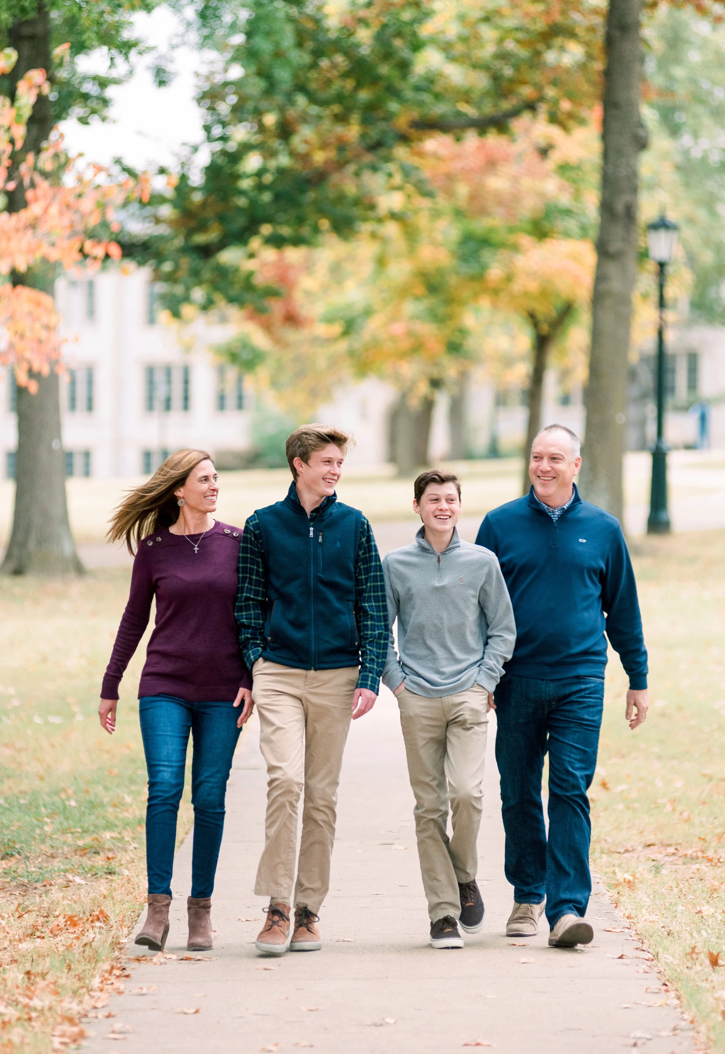 Family walking on a sidewalk in a park during fall, smiling and enjoying the outdoors with colorful autumn trees in the background.