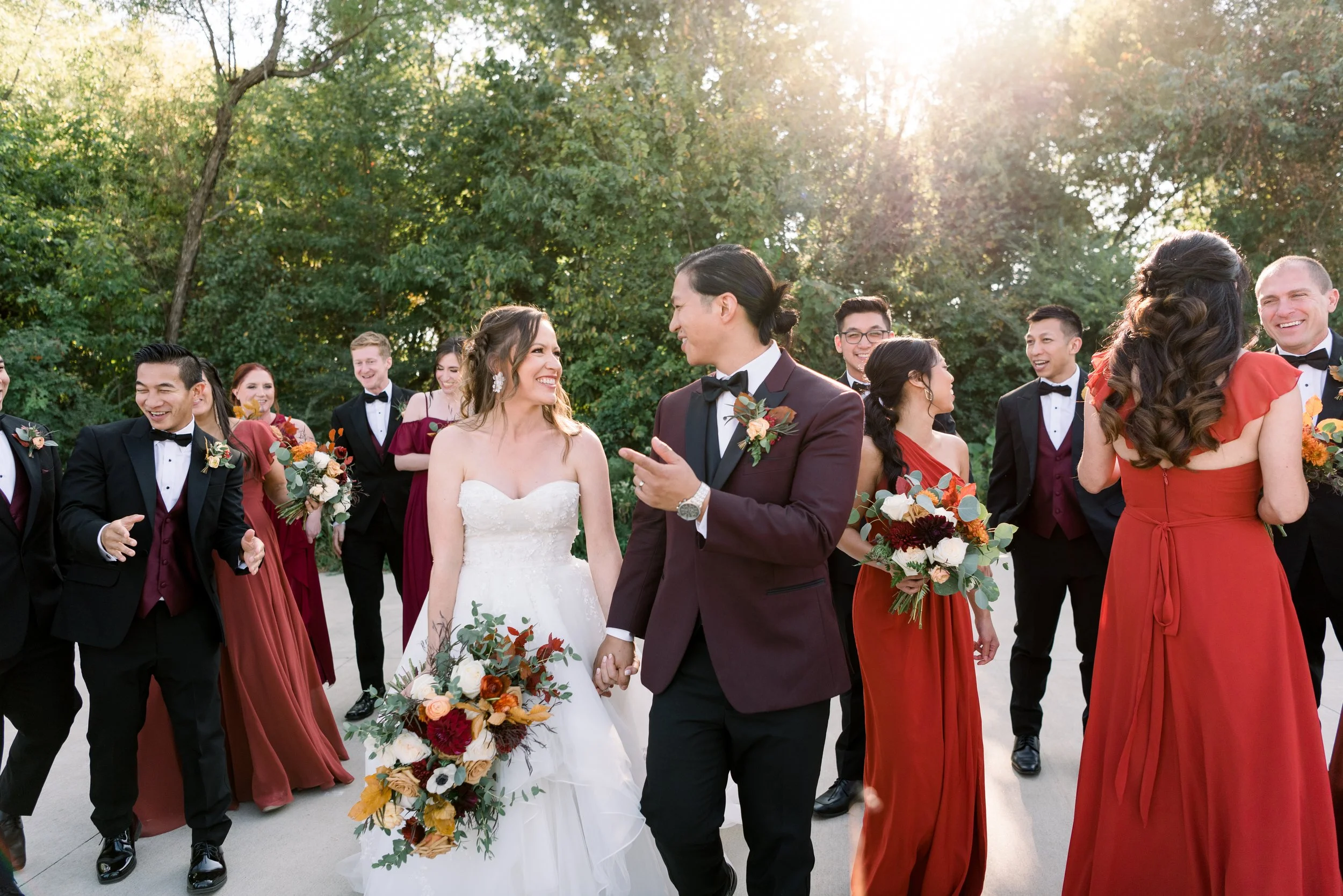 A wedding celebration outdoors with a bride and groom holding hands and smiling, surrounded by bridesmaids and groomsmen wearing formal attire, with trees and sunlight in the background.