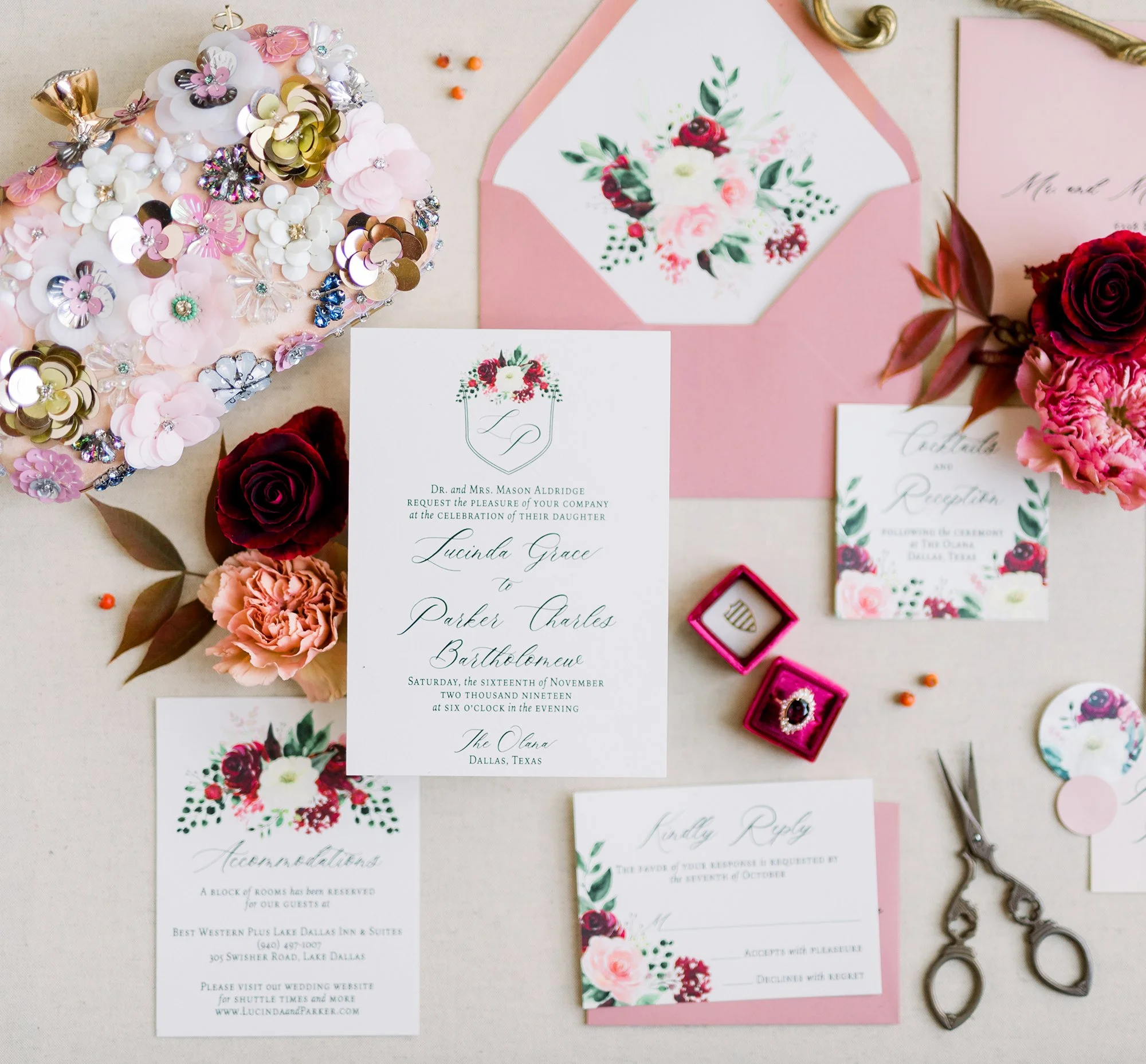 Wedding invitation suite with floral and greenery designs, pink envelope, bouquet of flowers, rhinestone-studded heart decoration, pink jewelry box with ring, and scissors on a white surface.