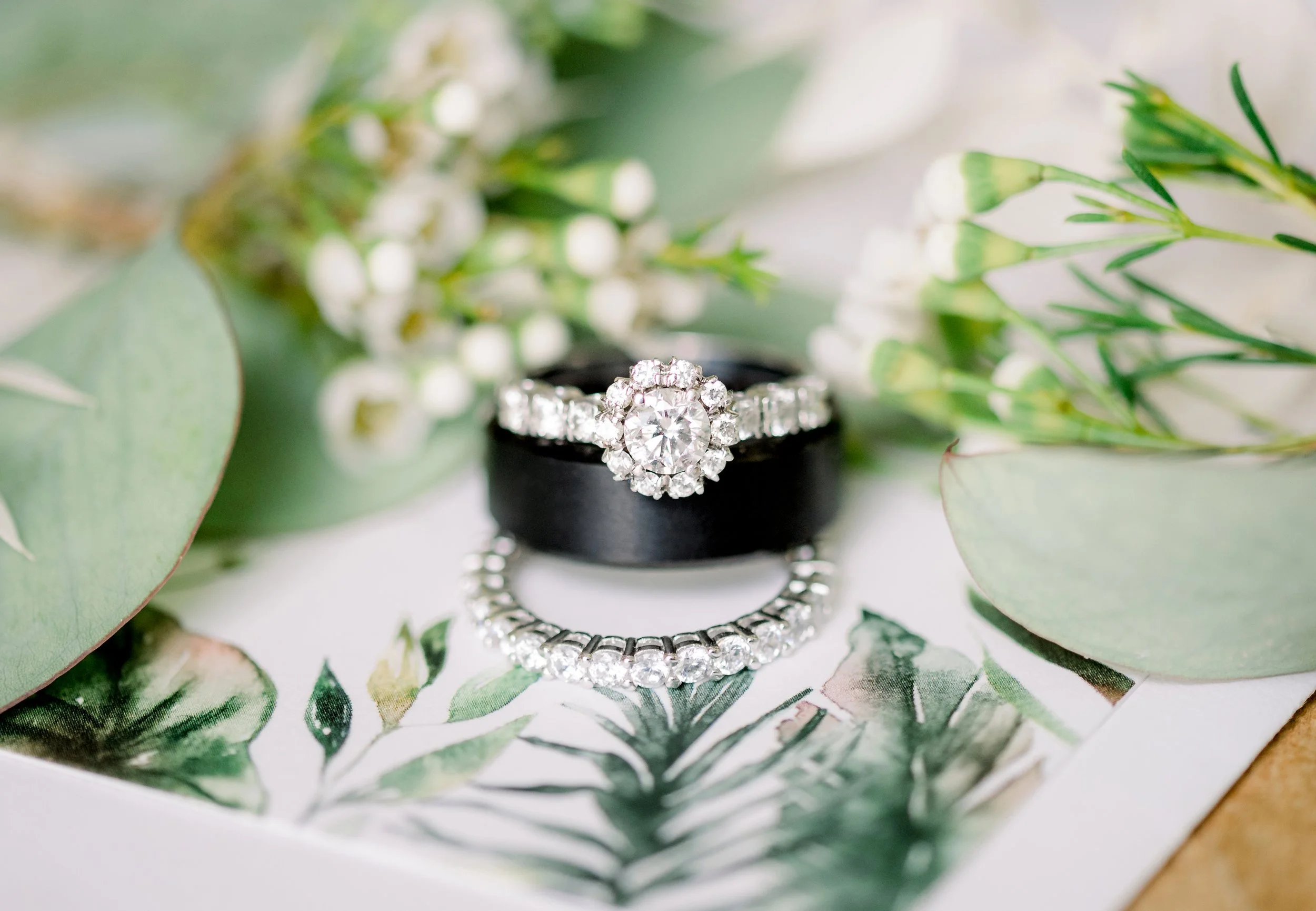A diamond engagement ring and a matching diamond wedding band are displayed on a black ring holder, surrounded by green leaves and white flowers on a botanical print background.