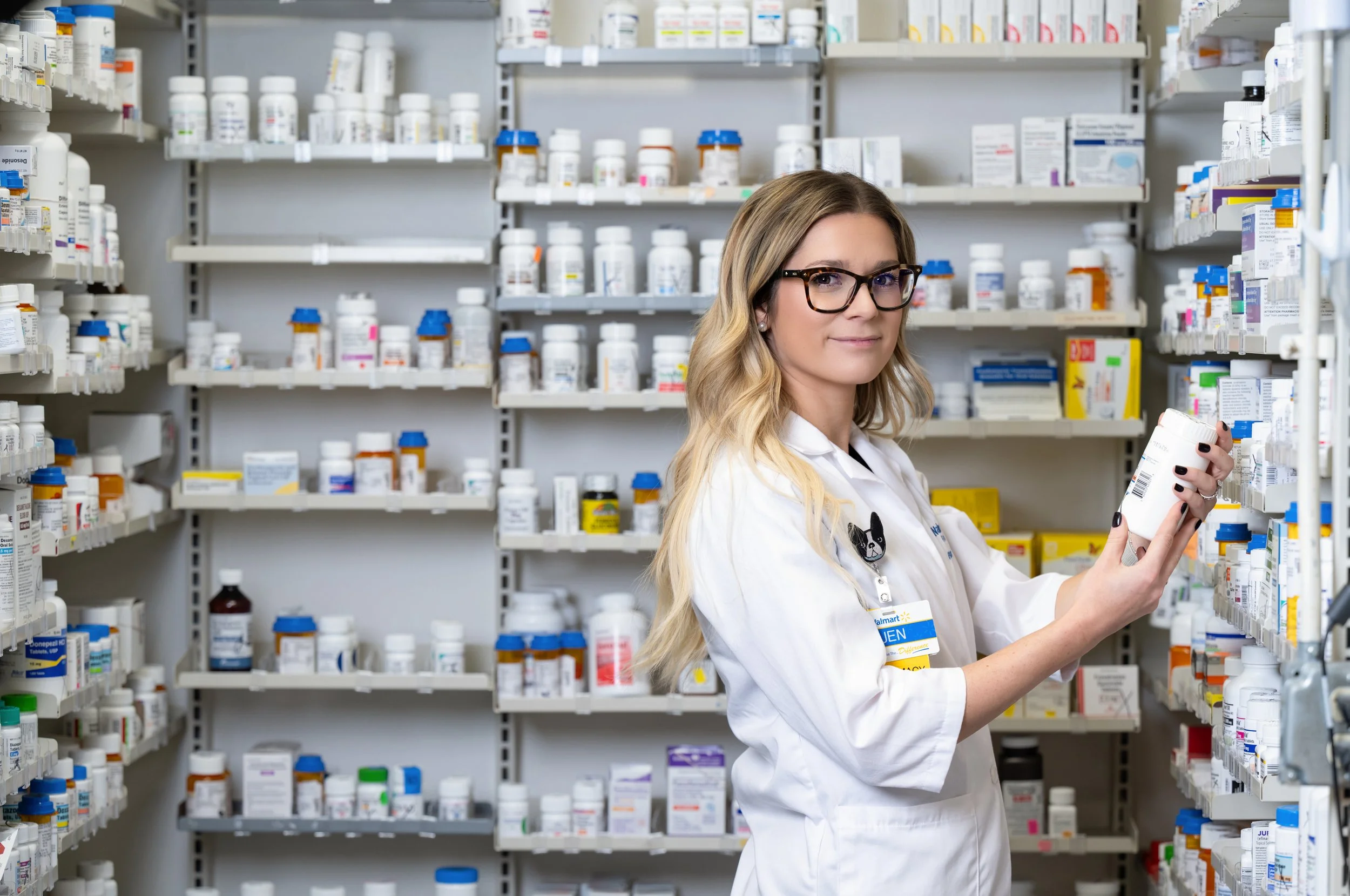 A female pharmacist wearing glasses and a white coat with a name tag, standing in an aisle of a pharmacy surrounded by shelves filled with medication bottles and boxes.