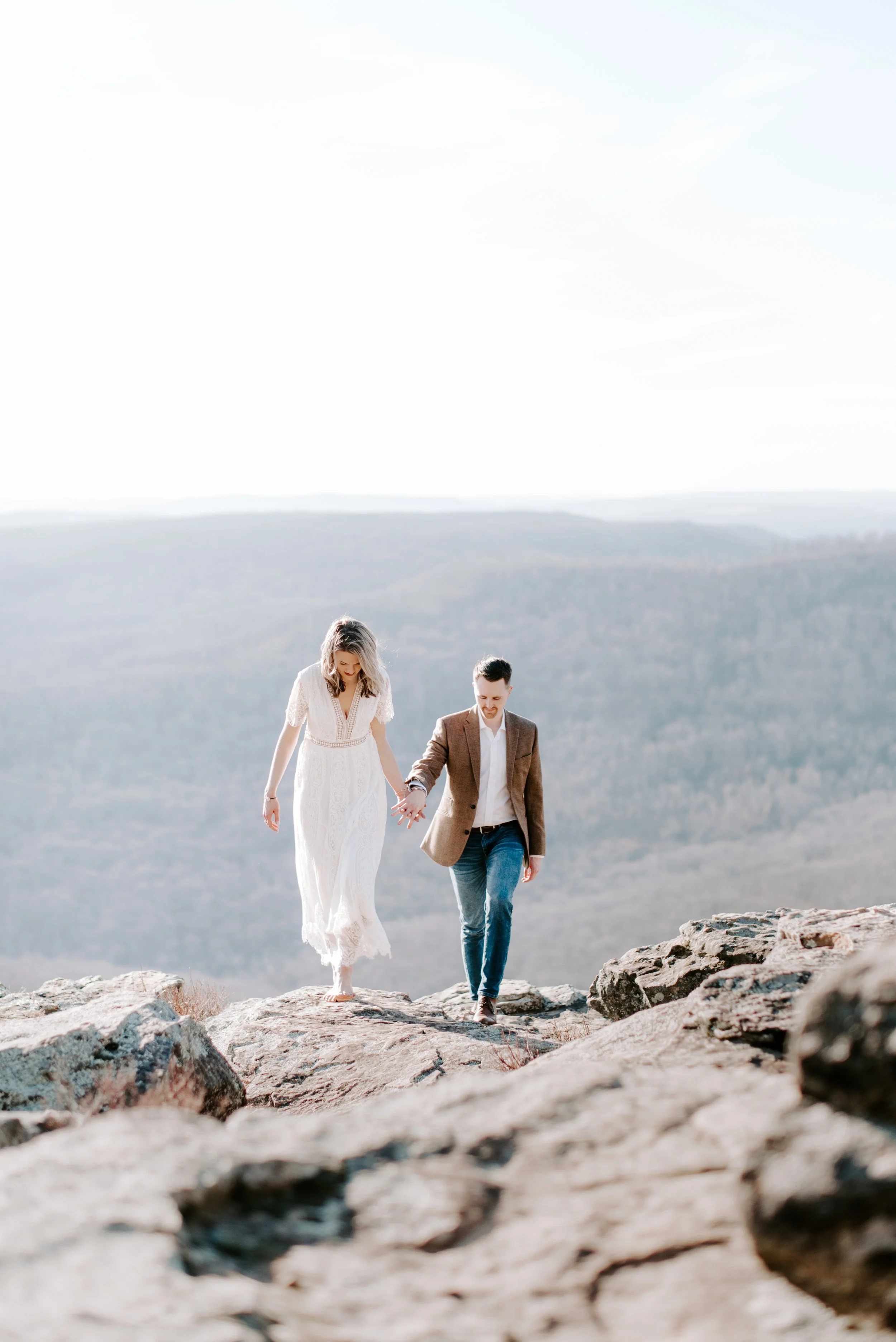 A couple walking hand in hand on rocky terrain in a scenic mountainous landscape, with a clear sky and distant hills.