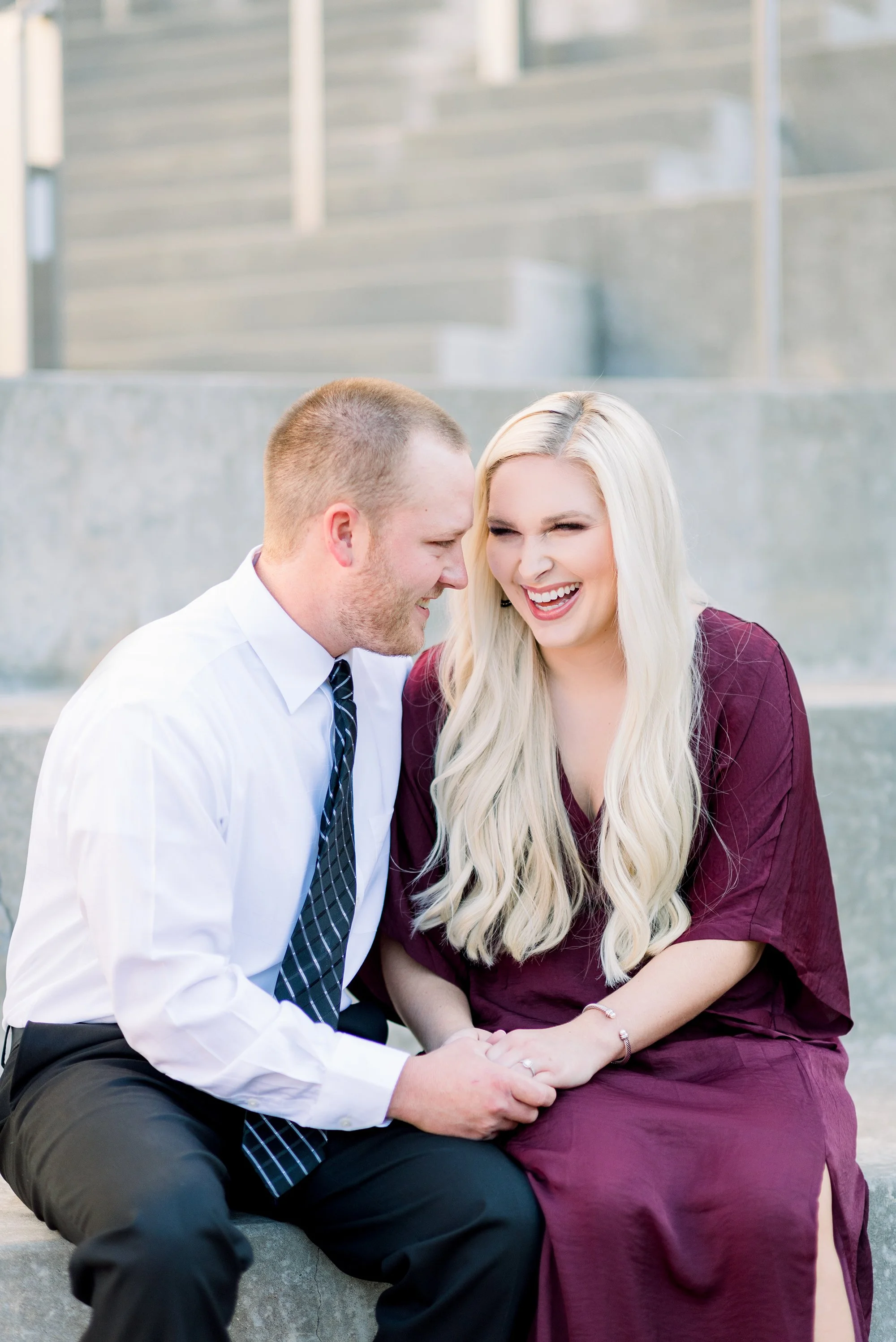 A happy couple is sitting together on concrete steps, smiling and laughing. The man has short hair, is dressed in a white shirt and black pants with a striped tie. The woman has long blonde hair, is wearing a maroon dress, and is showing her engageme