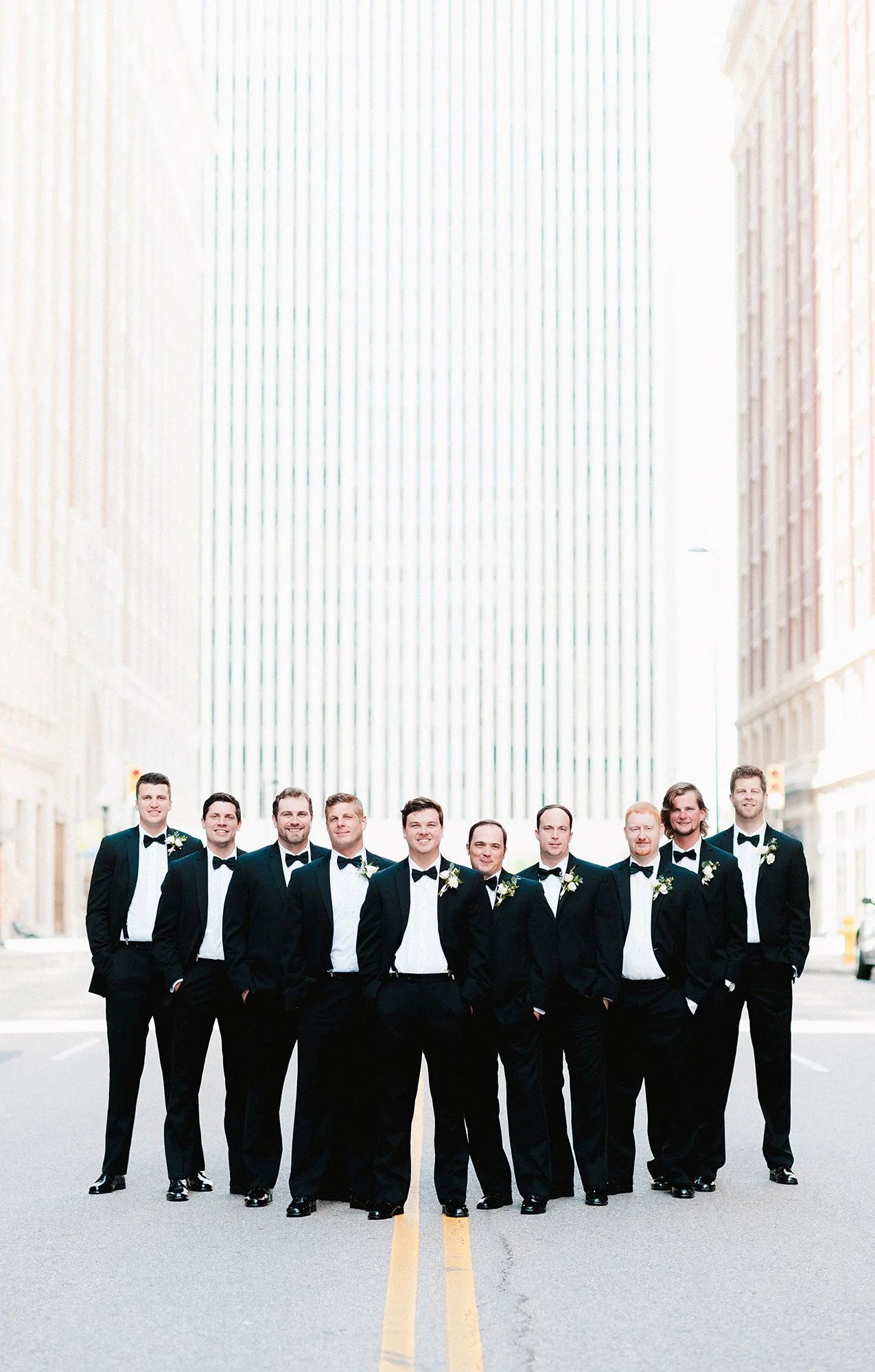 Group of men in tuxedos standing on city street.