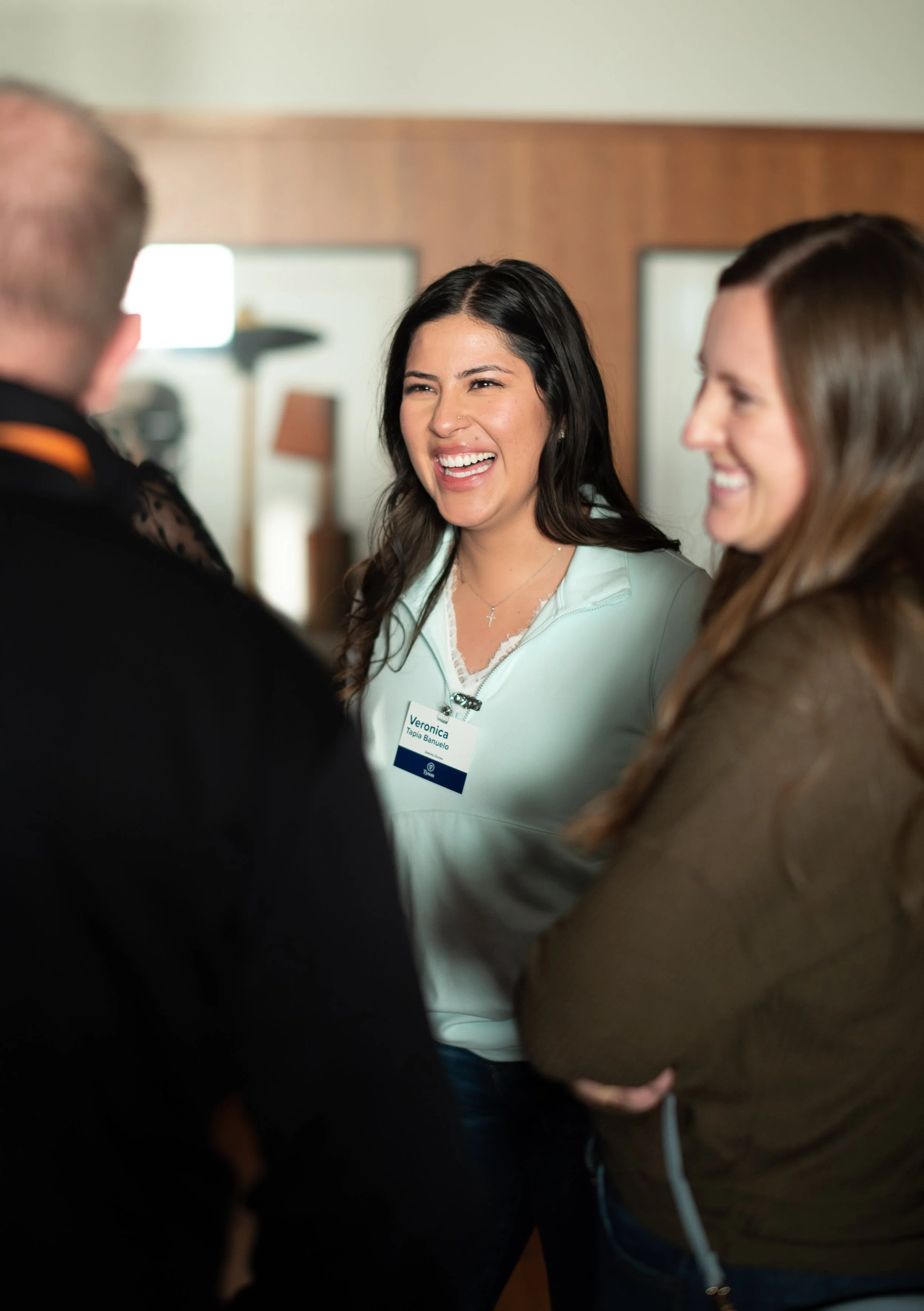 Three people engaged in conversation, smiling, at a social or professional event, with a woman wearing a name tag.