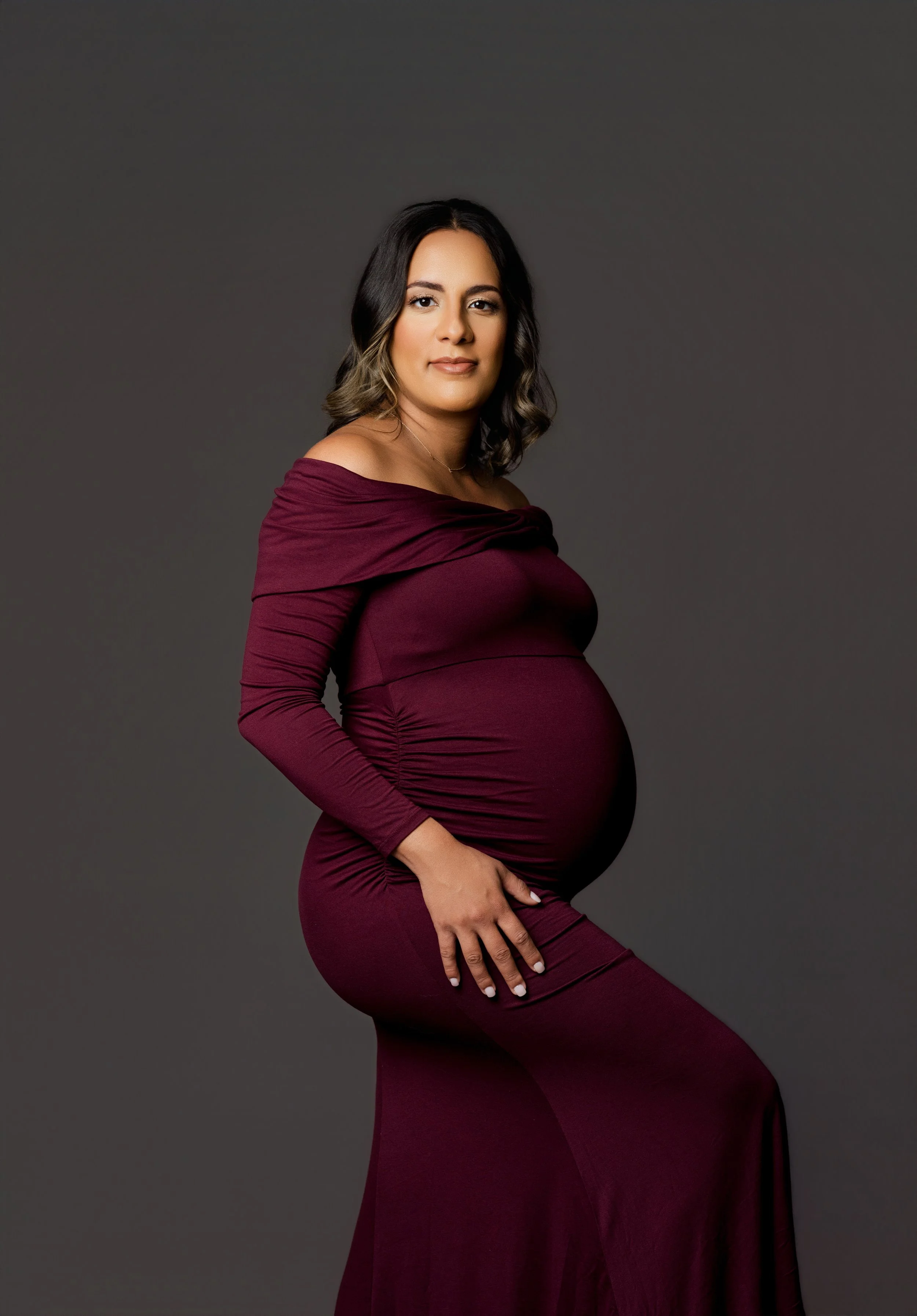 A pregnant woman with wavy dark hair in a maroon off-shoulder dress poses against a gray background.