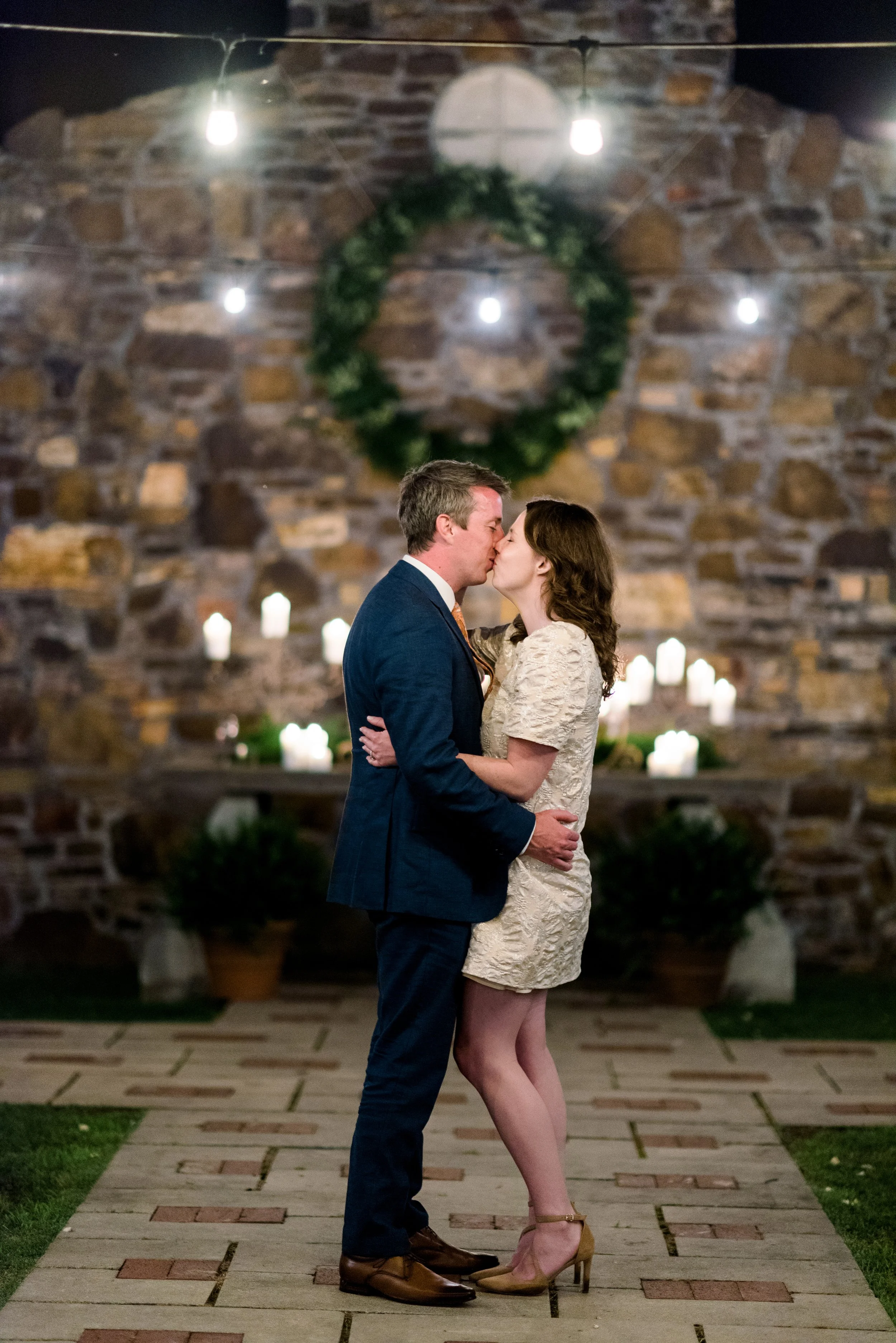A couple sharing a kiss at night in front of a stone wall decorated with a wreath and candles, with string lights overhead.
