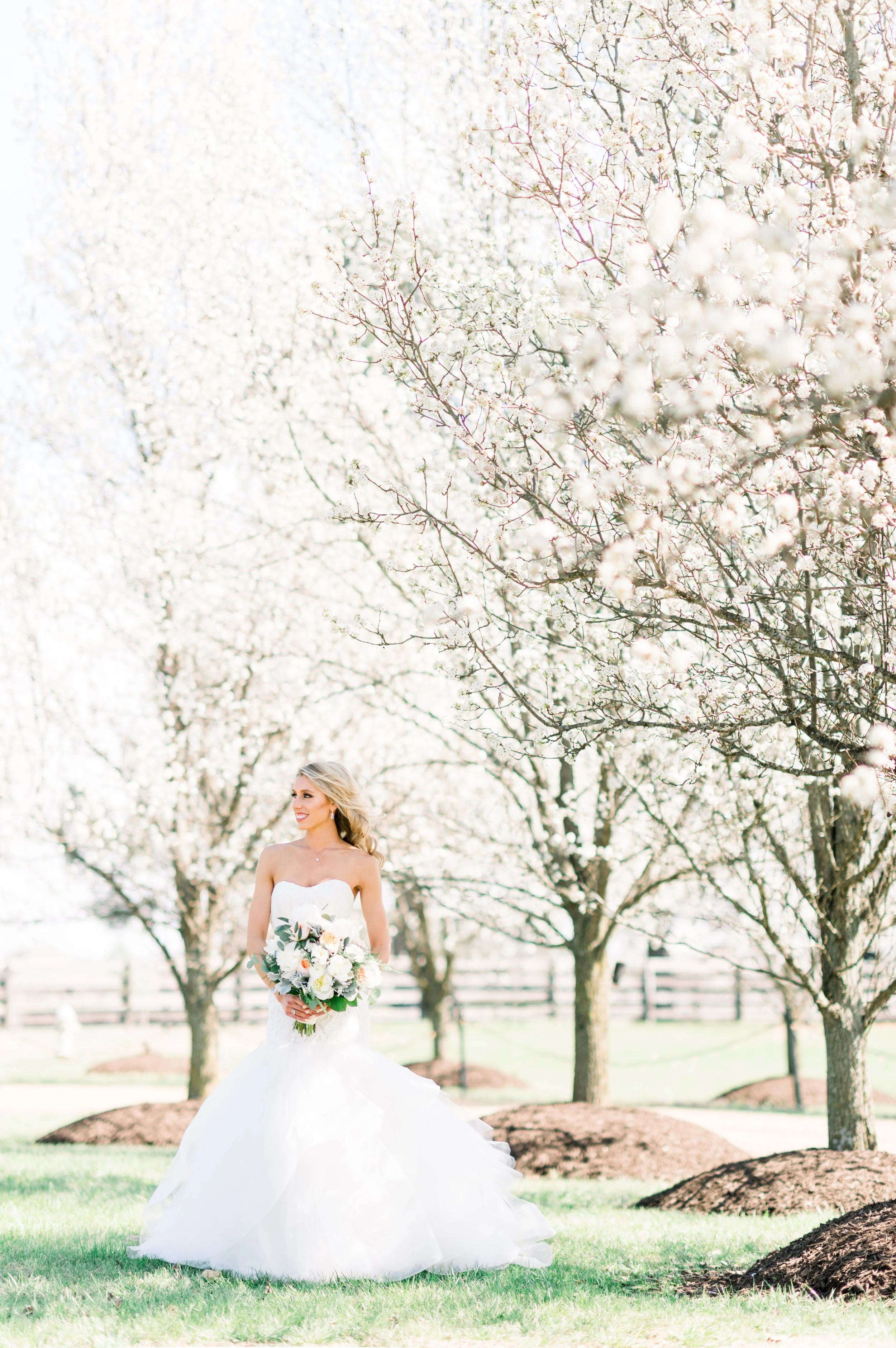 A bride in a white wedding dress holding a bouquet of flowers stands outdoors under blooming trees in spring.