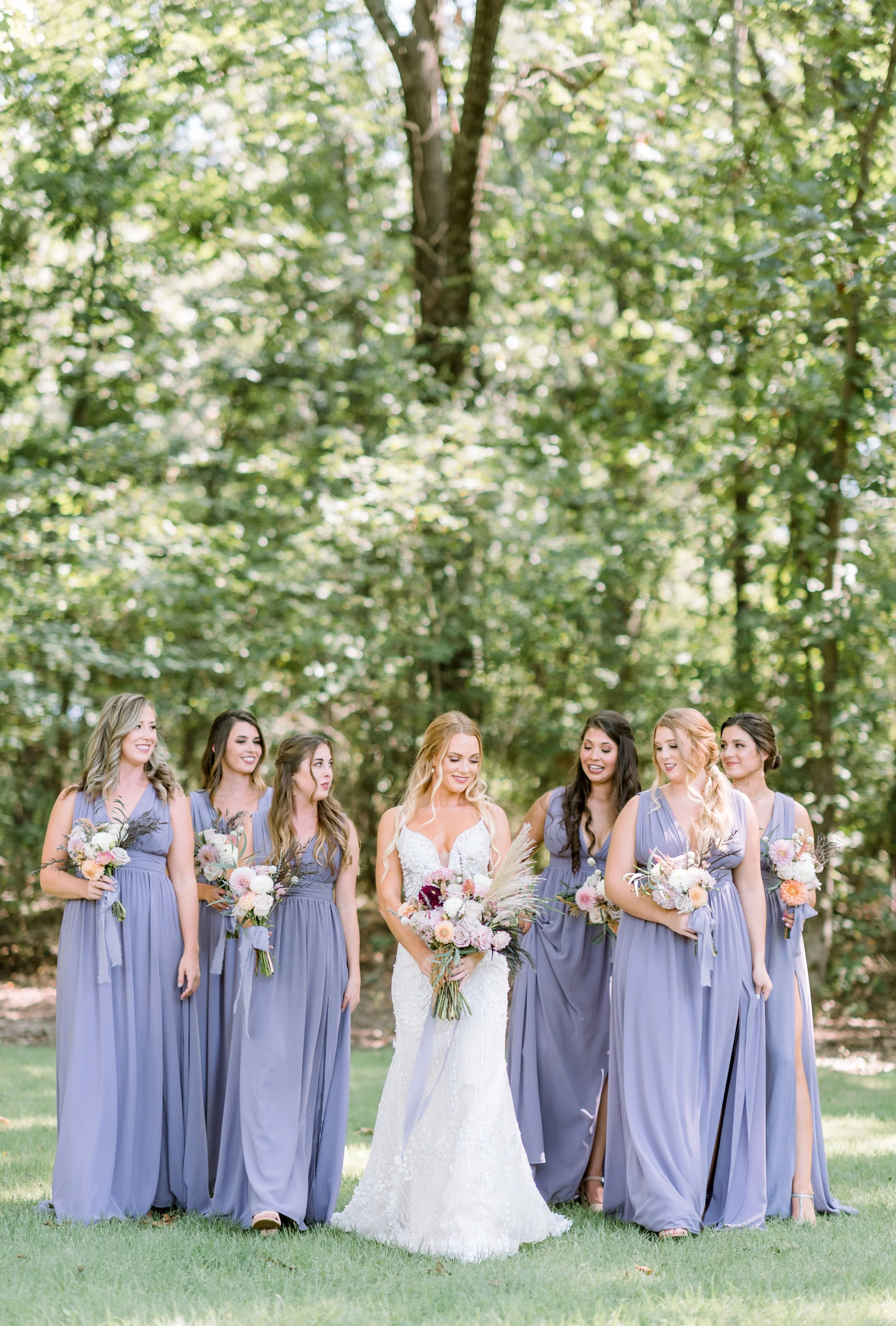 Bride in white wedding dress holding a bouquet of flowers surrounded by six bridesmaids in lavender dresses holding bouquets, outdoors in a wooded area.