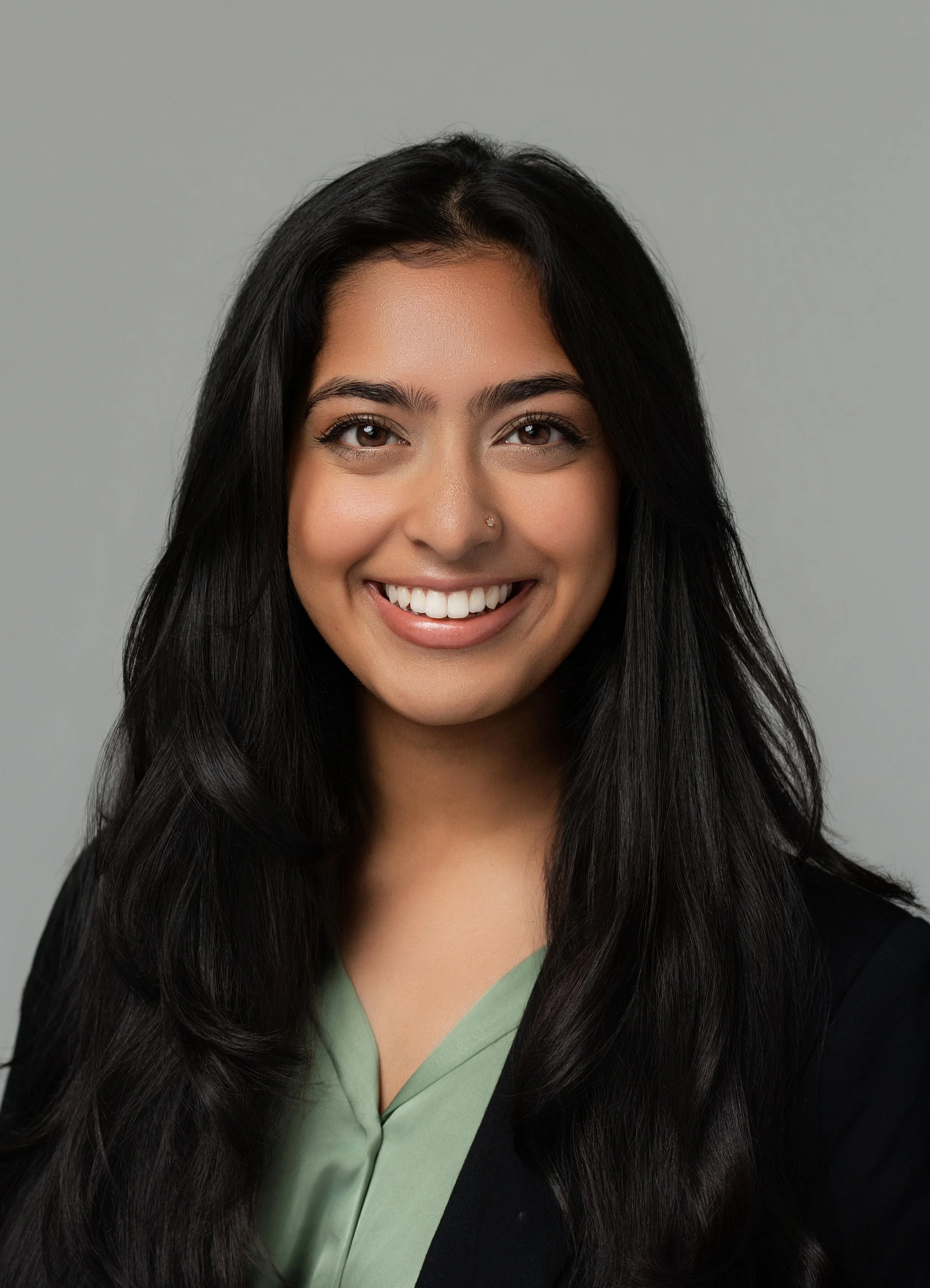 A smiling woman with long black hair, wearing a black blazer over a green top, against a plain gray background.