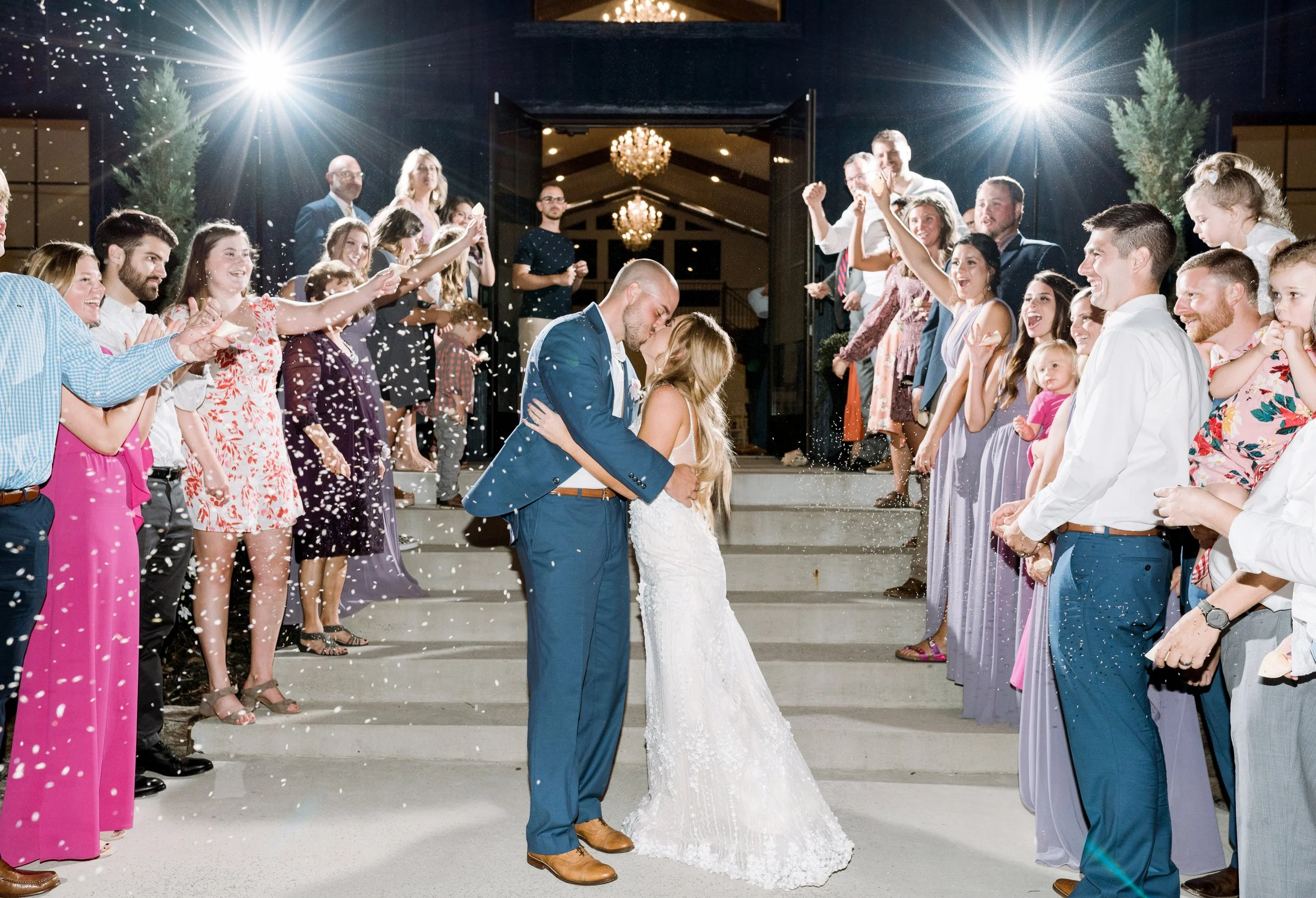 A couple dancing on the steps of a building at night, surrounded by cheering guests throwing confetti, at a wedding celebration.
