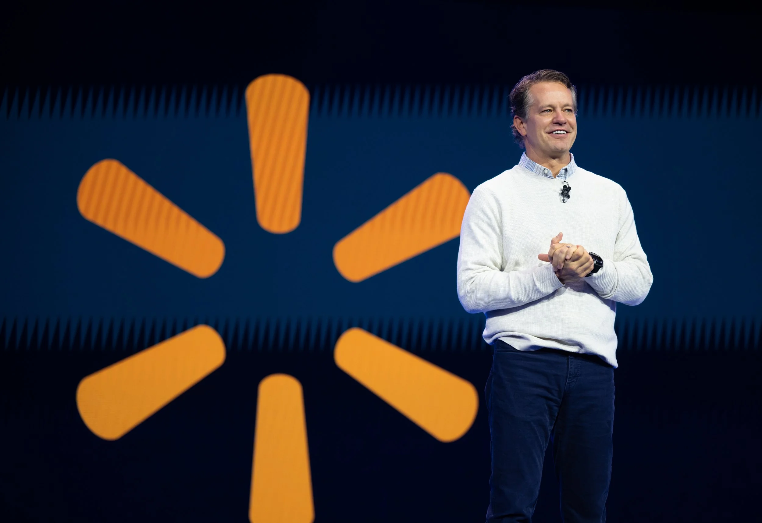 A man standing on stage, smiling, wearing a white sweater and dark pants, with a large Walmart logo in the background.