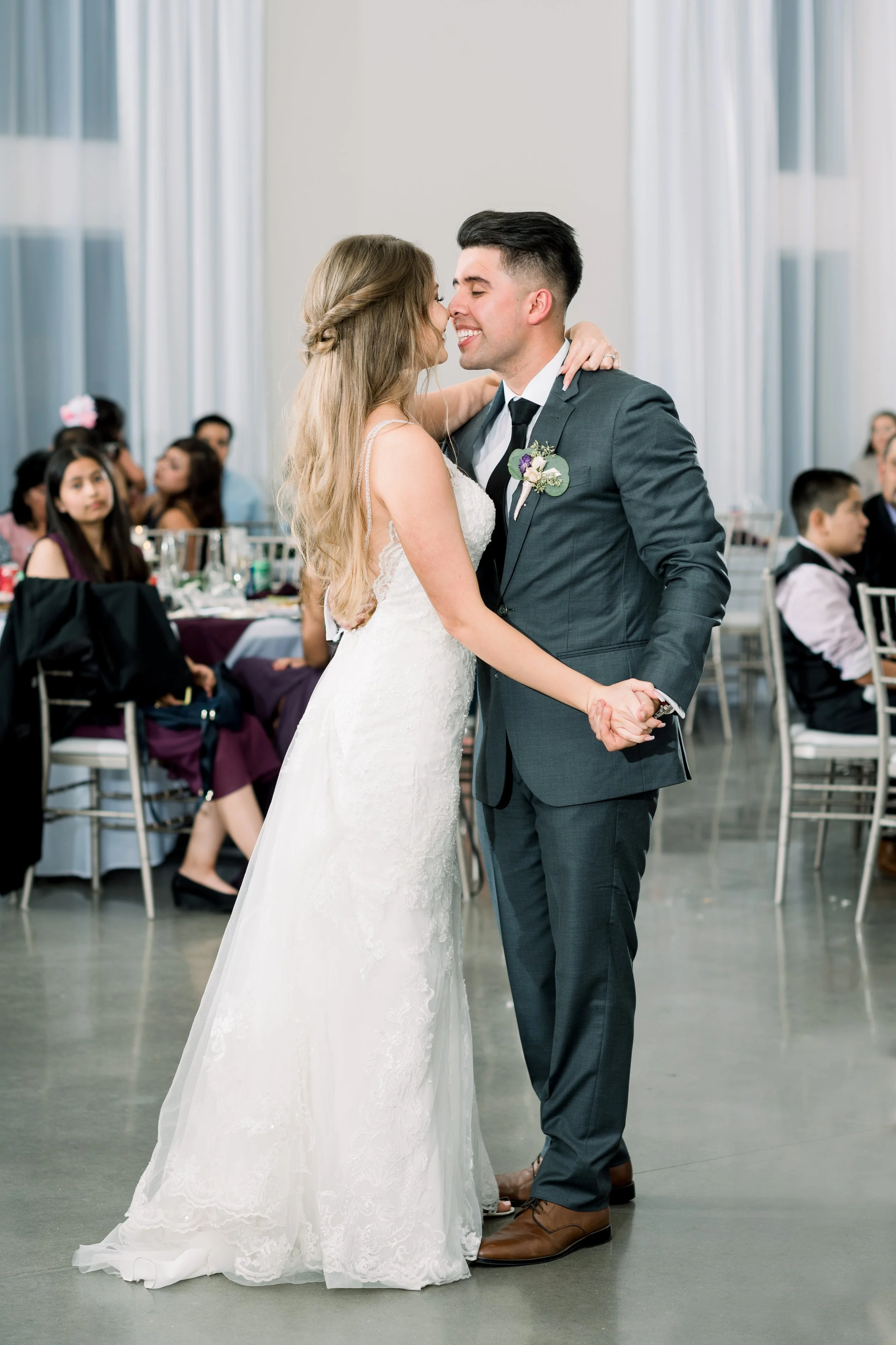 A bride and groom share a dance at their wedding reception indoors, with guests seated at tables in the background.