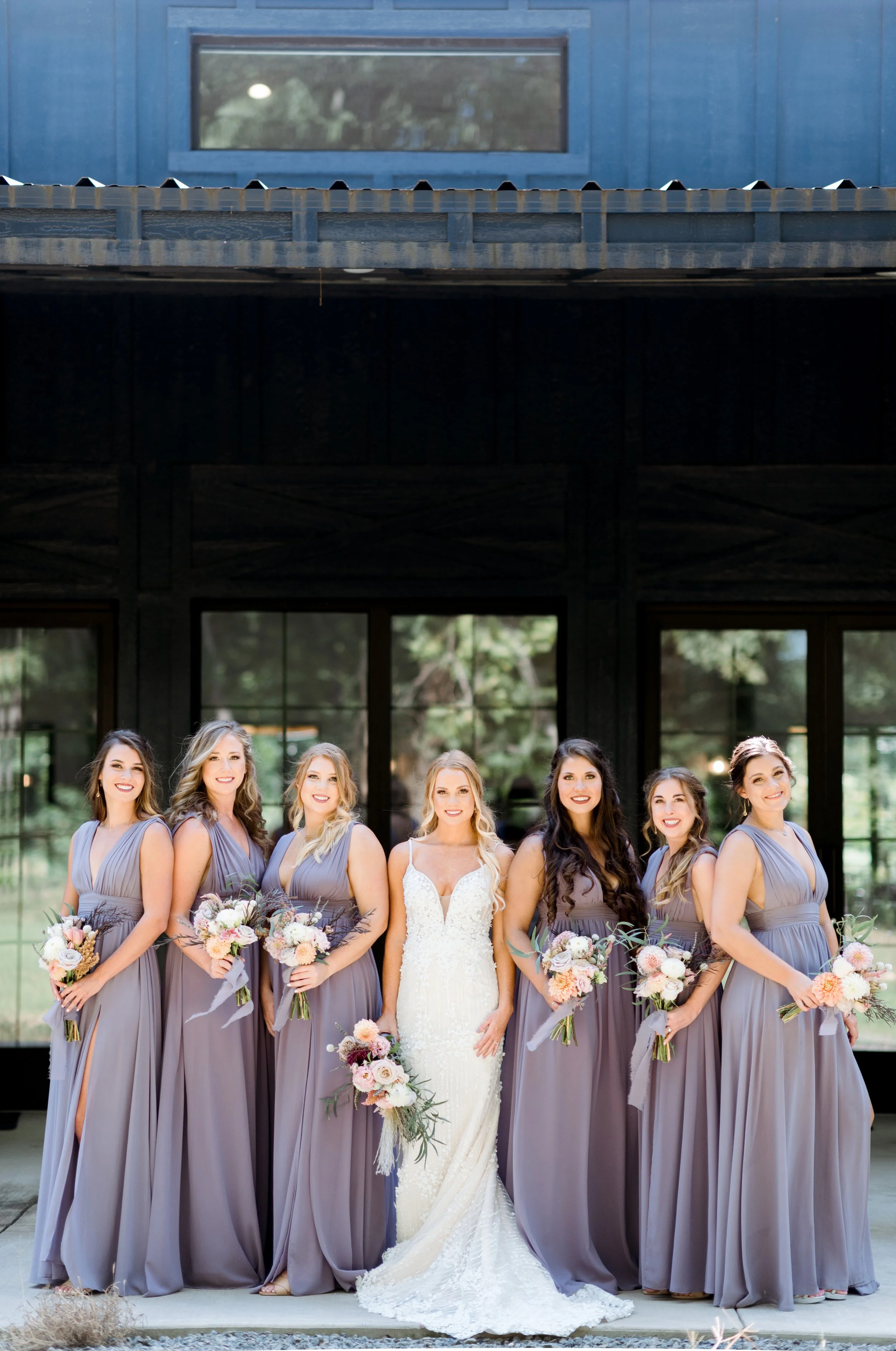 A bride in a white wedding gown standing with six bridesmaids in matching mauve gowns, holding bouquets, outside in front of a dark modern building with large windows.