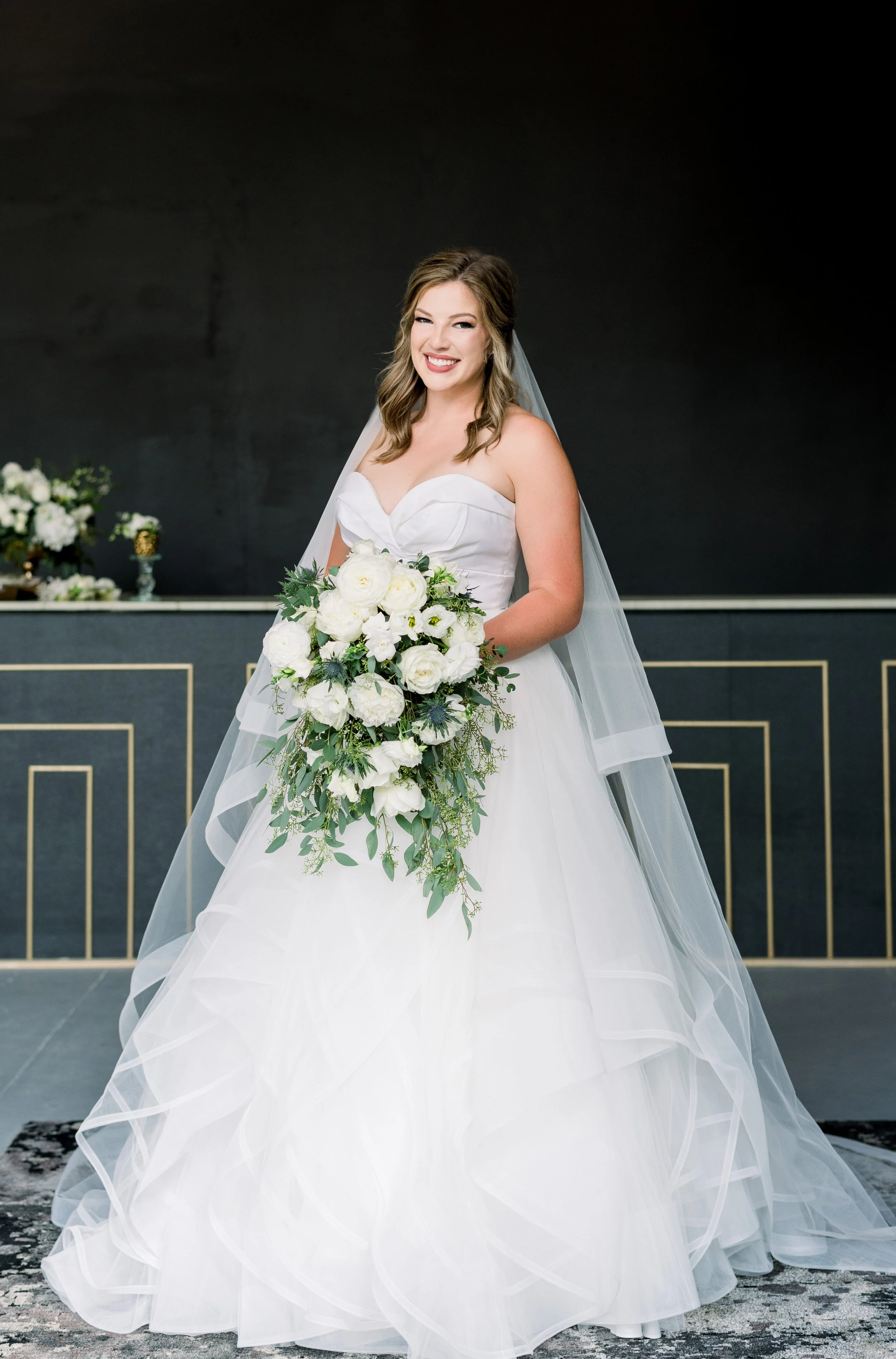 A smiling bride in a white wedding gown holding a large cascading bouquet of white roses and greenery.