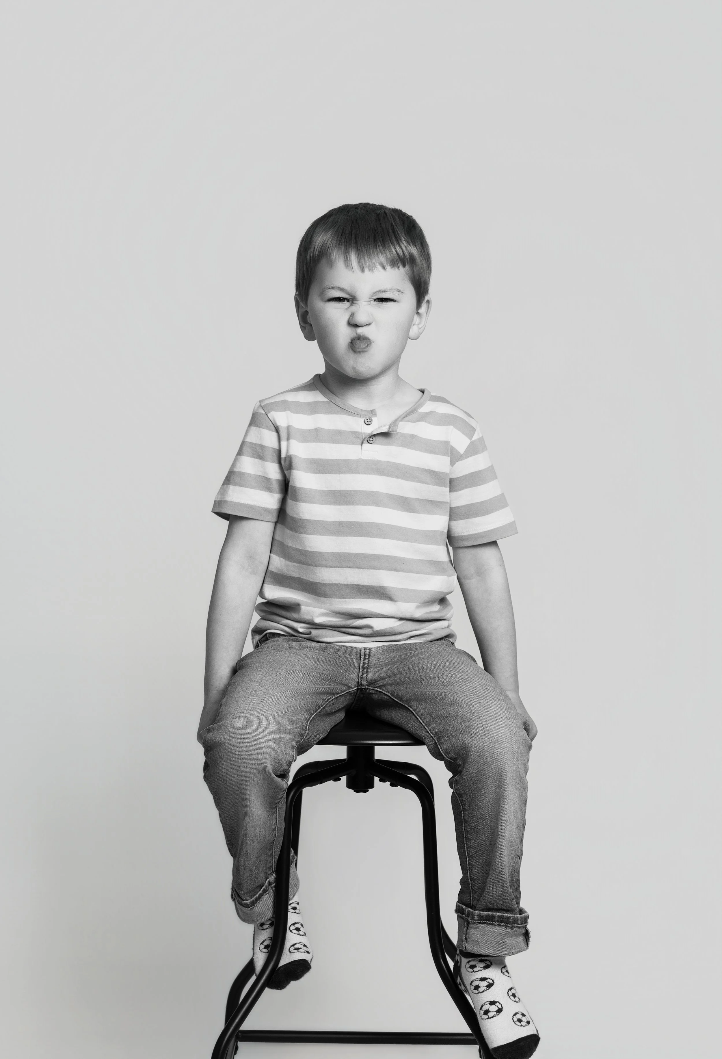 Black and white photo of a young boy sitting on a stool, making a funny face with pursed lips and squinted eyes, wearing a striped t-shirt, jeans, and soccer socks.