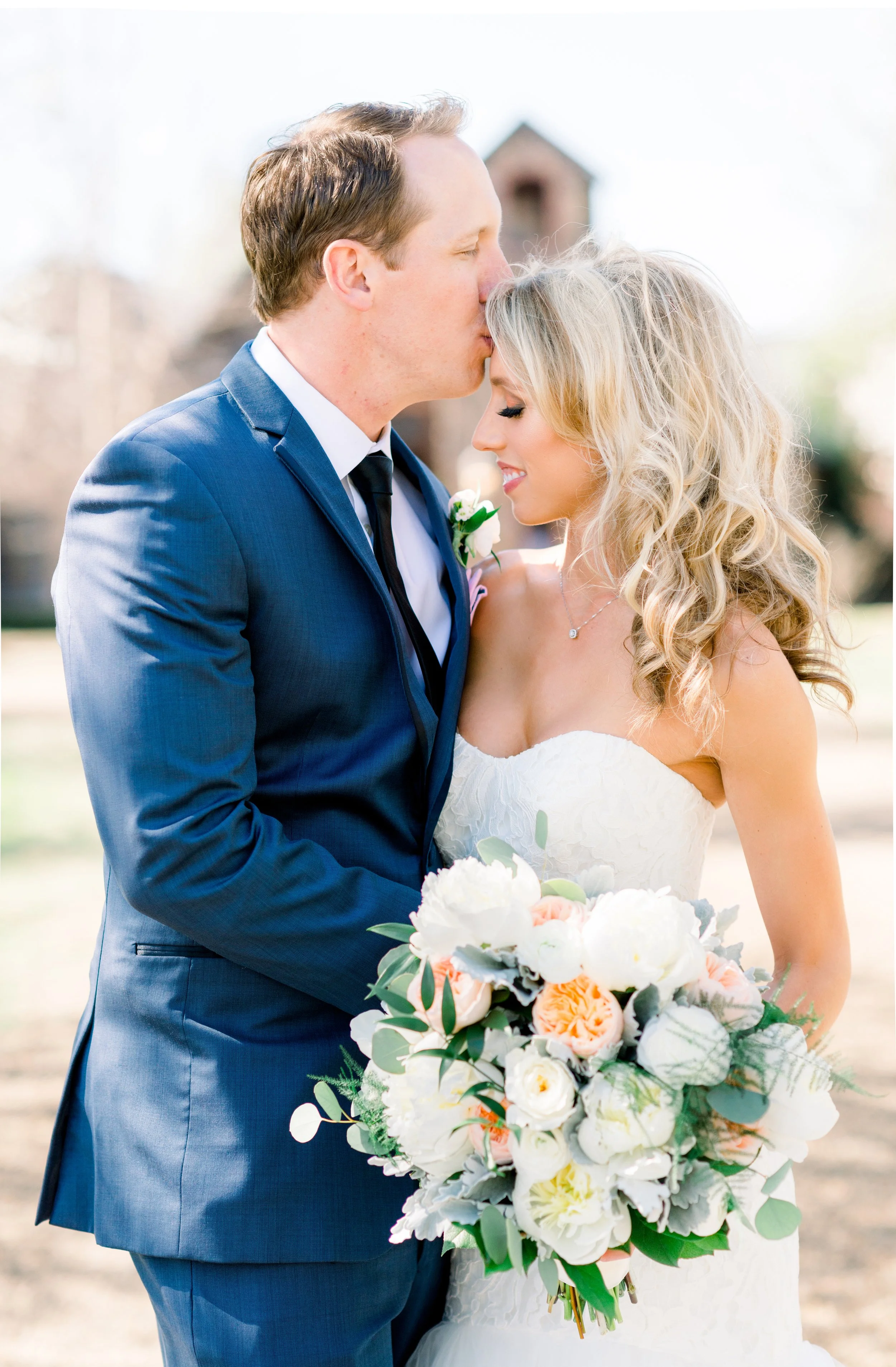 A bride and groom share a tender moment outdoors, with the groom kissing the bride's forehead, she holding a bouquet of white and pastel flowers.