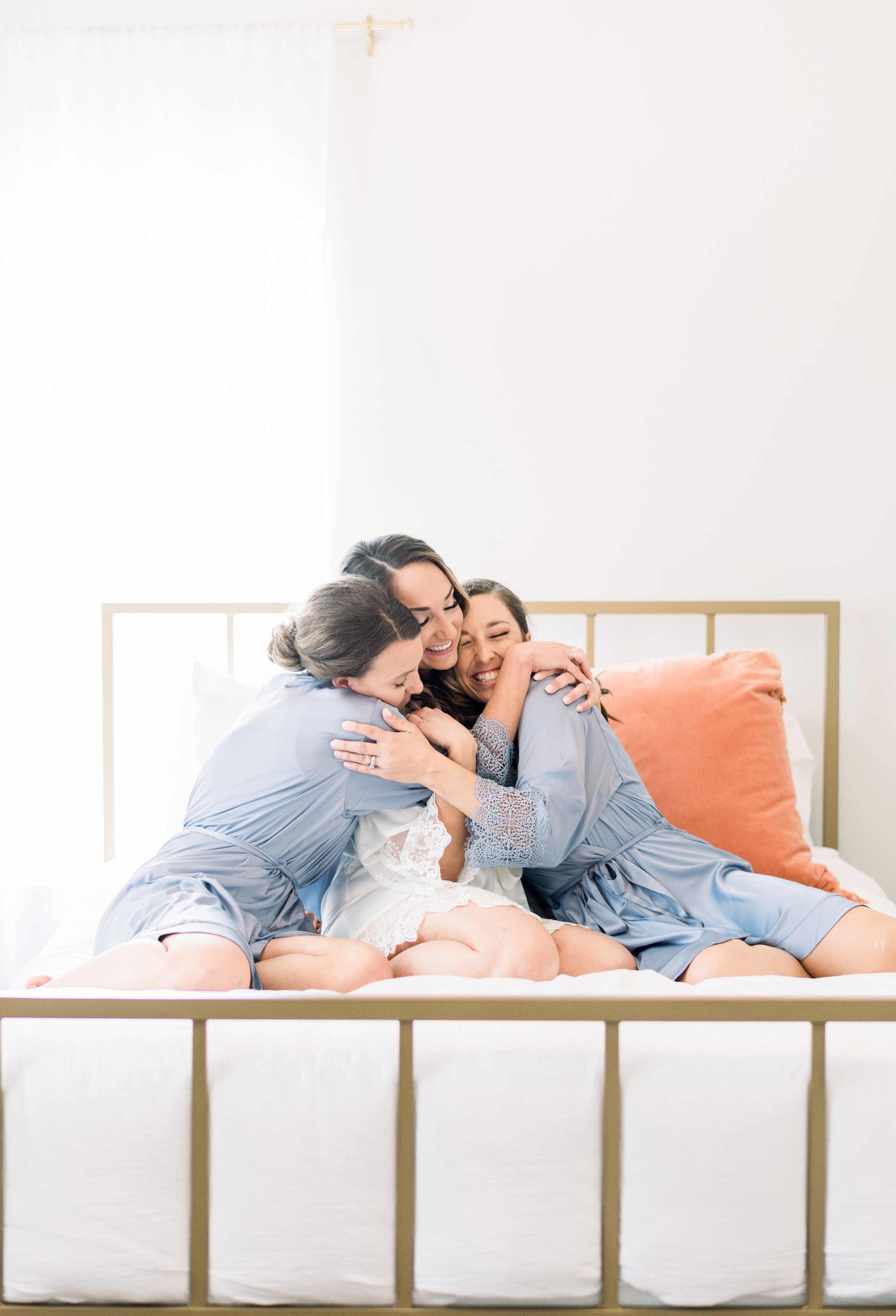 Three women in matching blue robes embracing and smiling on a bed with a white background and a orange pillow behind them.