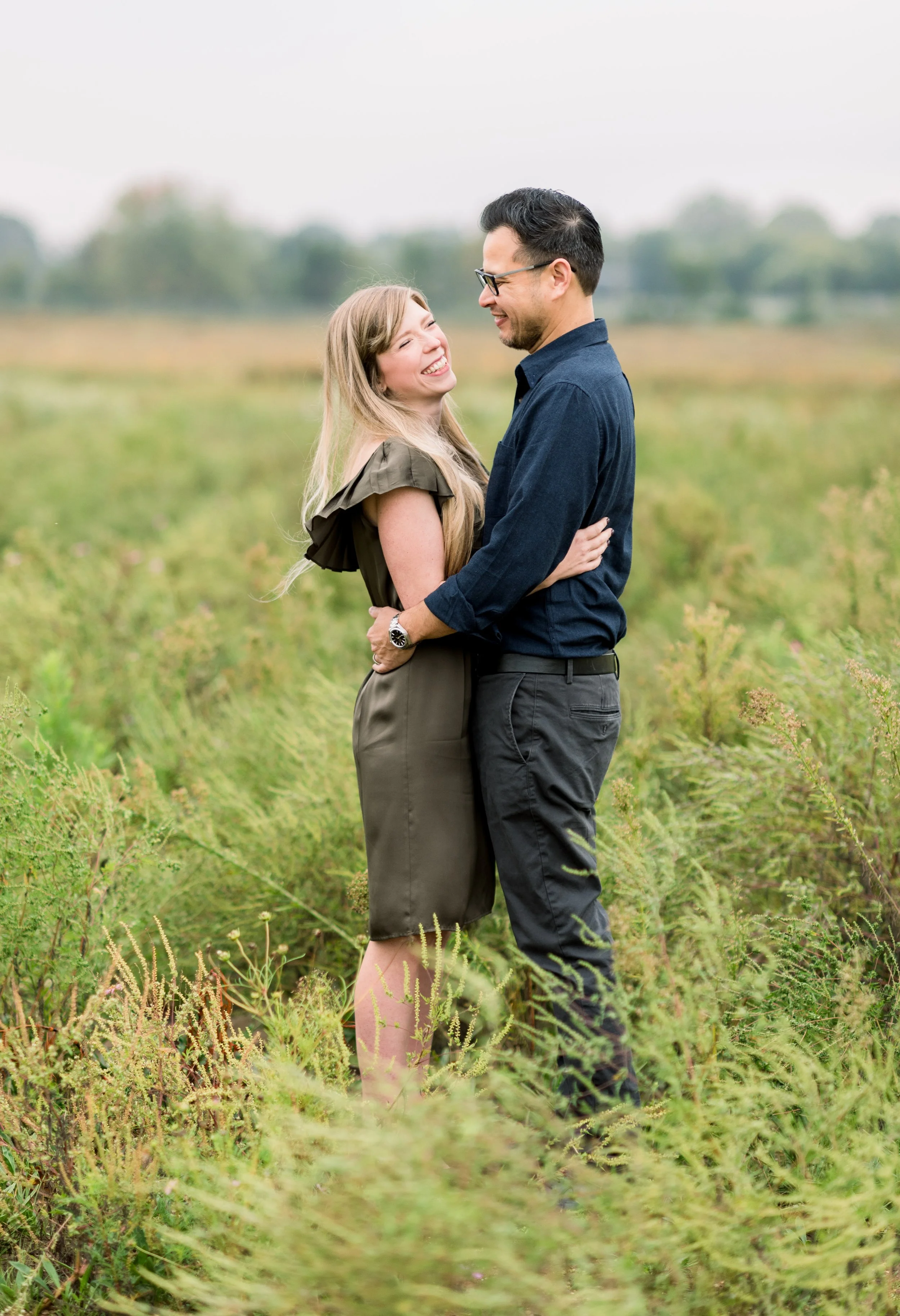 A happy couple stands in a grassy field, smiling and embracing each other on a cloudy day.