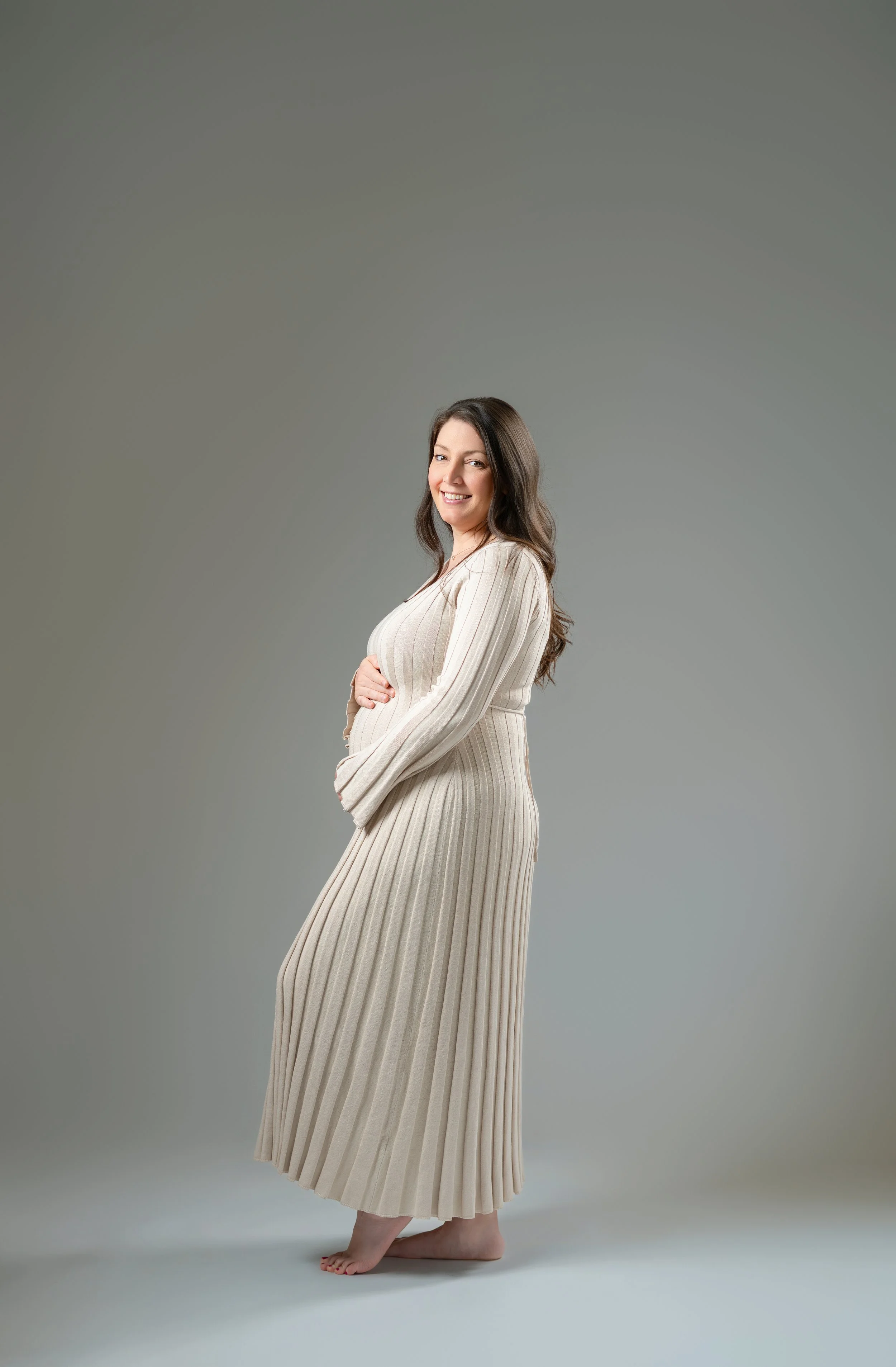 A pregnant woman in a beige, pleated dress, smiling, standing barefoot against a plain gray background.