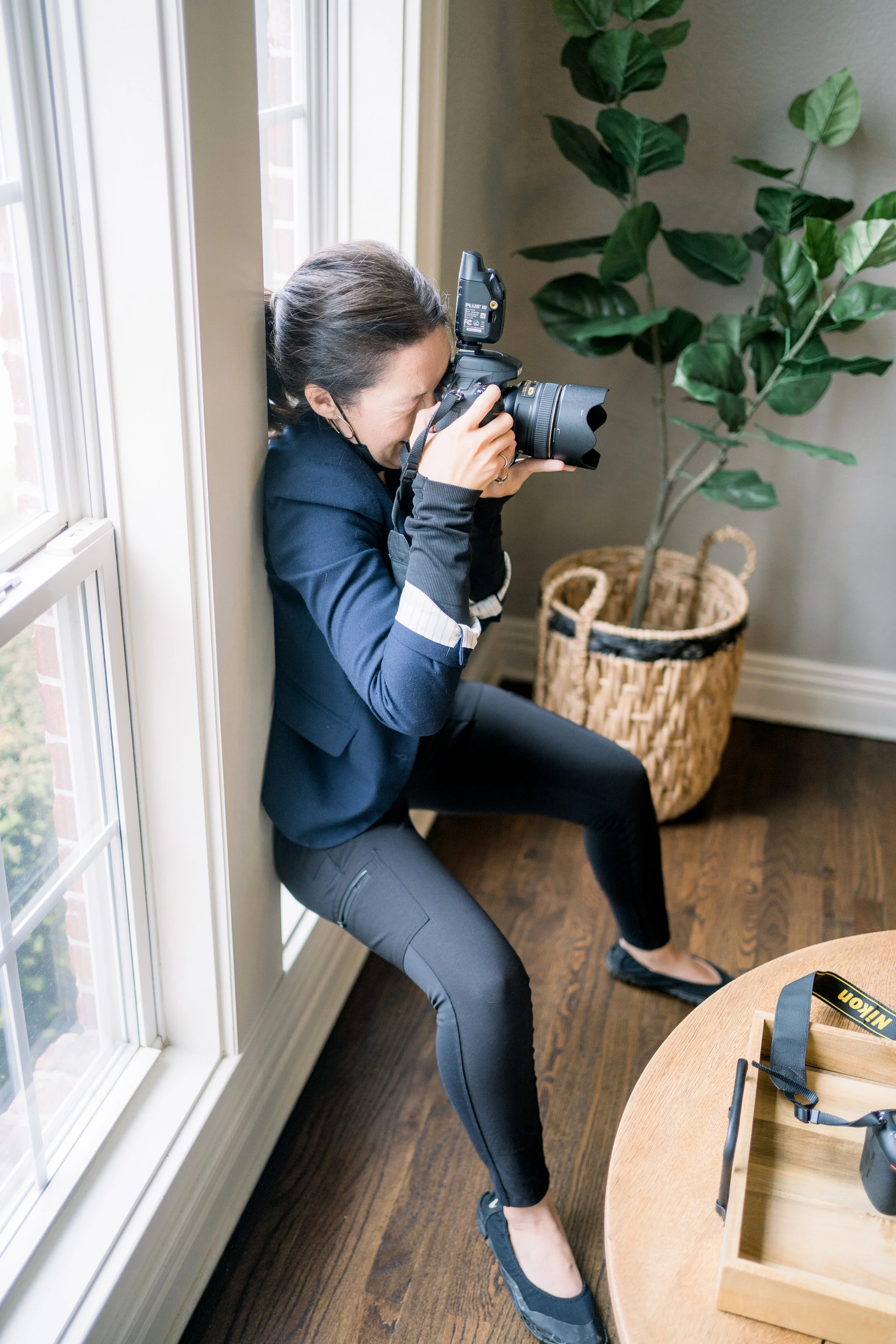 A woman taking a photograph with a professional camera while leaning against a wall near a window, with a large potted plant in the background.