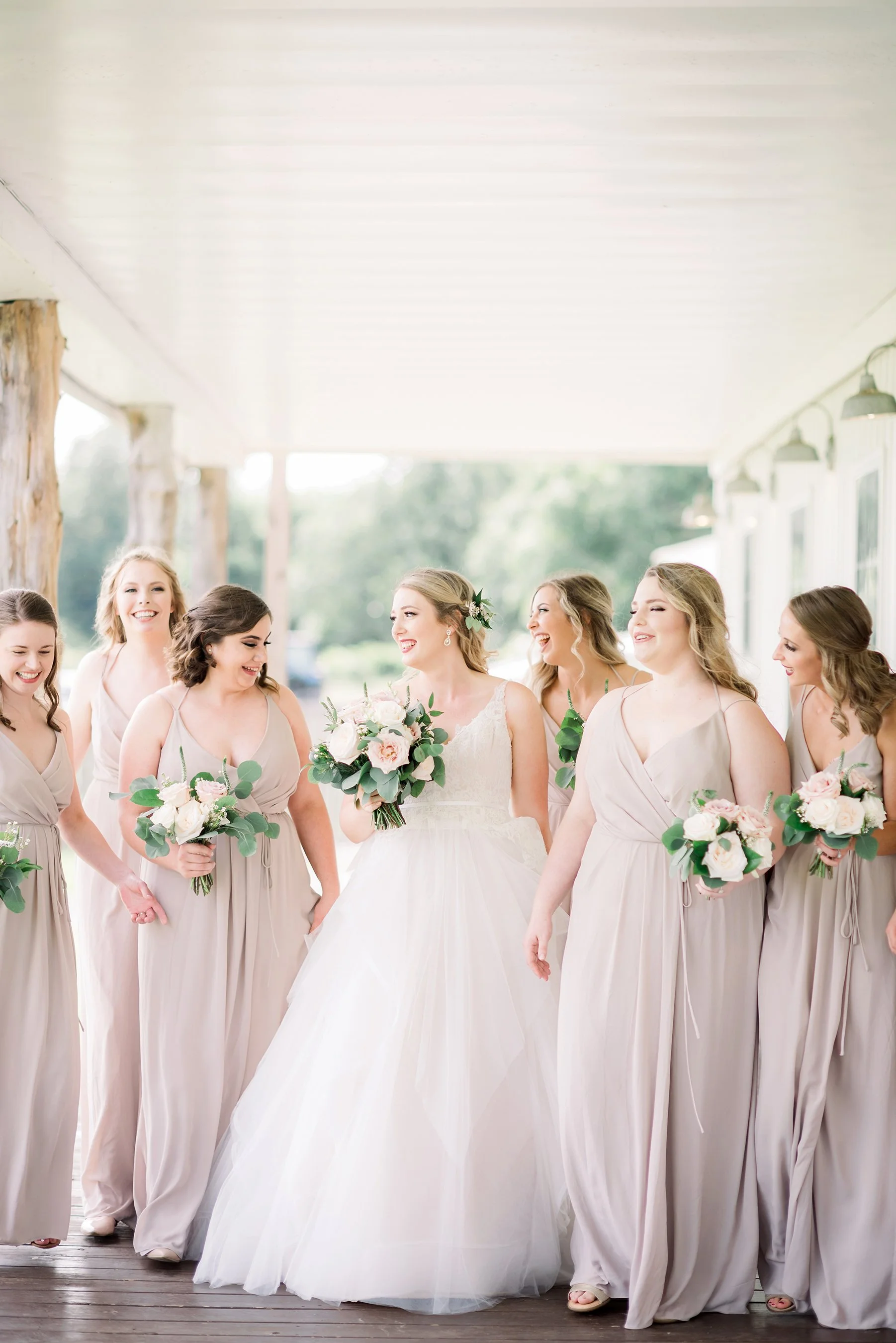 A bride and her bridesmaids walking and laughing together outdoors. The bride is holding a bouquet, wearing a white wedding gown. The bridesmaids are dressed in matching beige dresses, also holding bouquets.