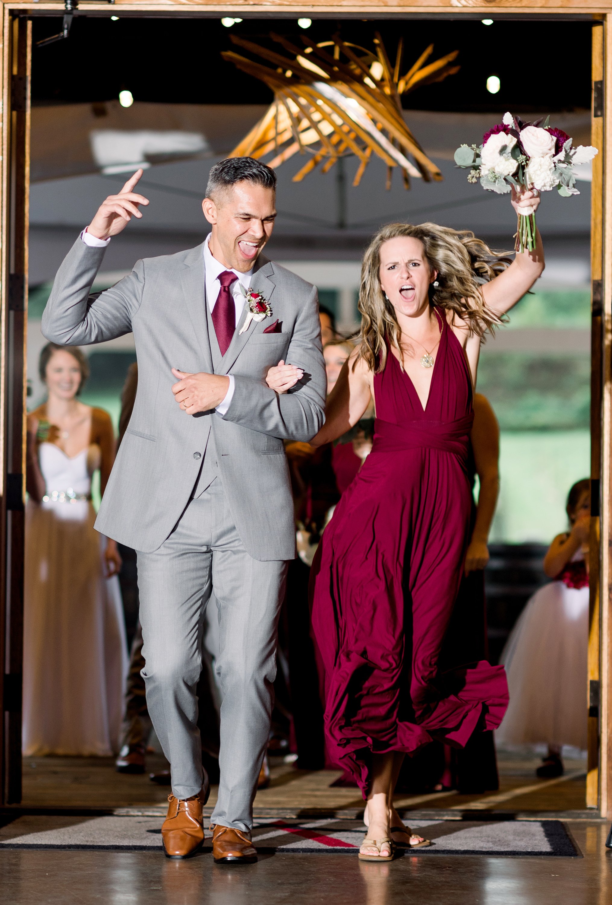 A wedding celebration with a man in a gray suit and a woman in a burgundy dress walking into a reception, smiling and raising her bouquet. Several bridesmaids and guests are visible in the background.