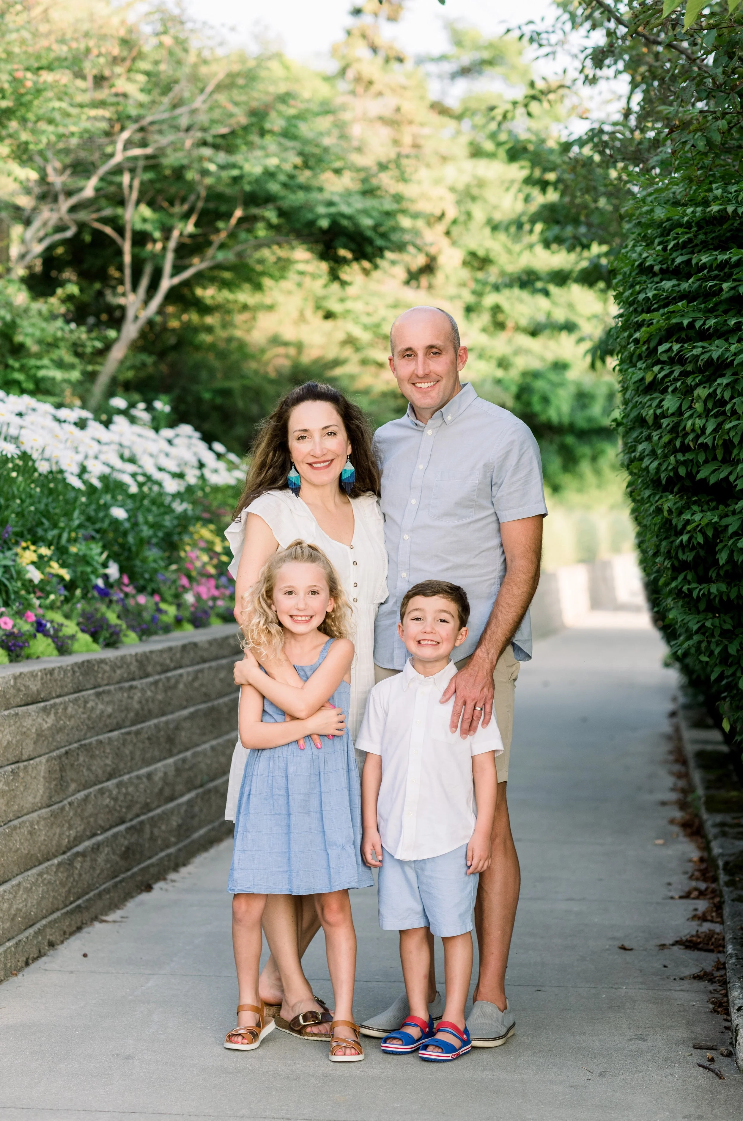 A happy family of five standing outdoors on a sidewalk, with a garden and trees in the background. The group includes a woman with long dark hair, a man, a young girl with curly blonde hair, and a young boy. All are smiling and dressed in casual summ