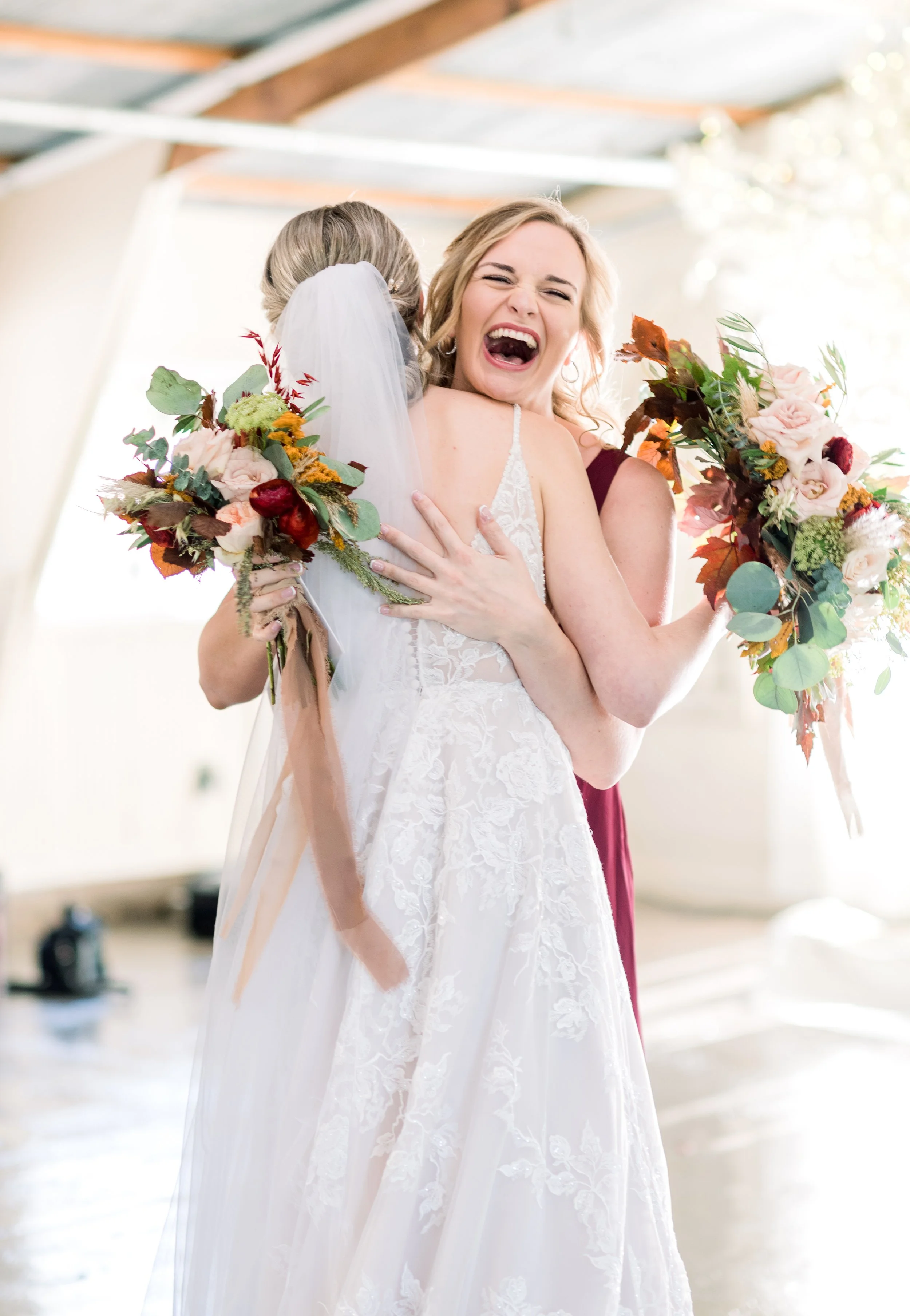 A bride in a white lace wedding gown and a woman in a burgundy dress embrace, holding bouquets of flowers. The bride's back is to the camera, and she has a veil. The woman is smiling with her eyes closed, joyfully hugging the bride.