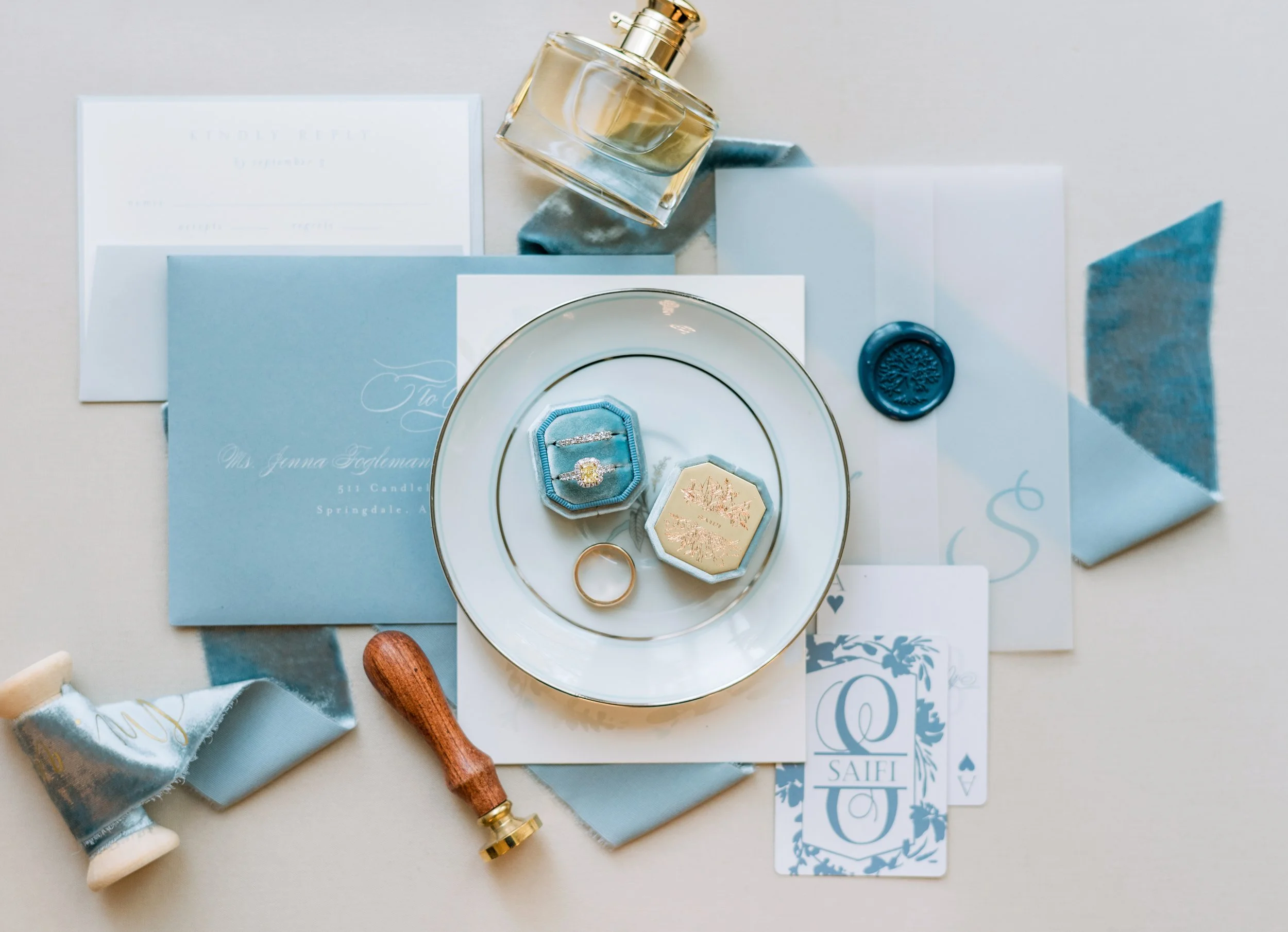 Flat lay of wedding invitation cards, blue and white envelopes, and a blue wax seal with a tree design, along with a glass bottle of perfume, a gold ring, a small blue ring box, a spool of blue ribbon, and a postage stamp featuring a wildlife illustr