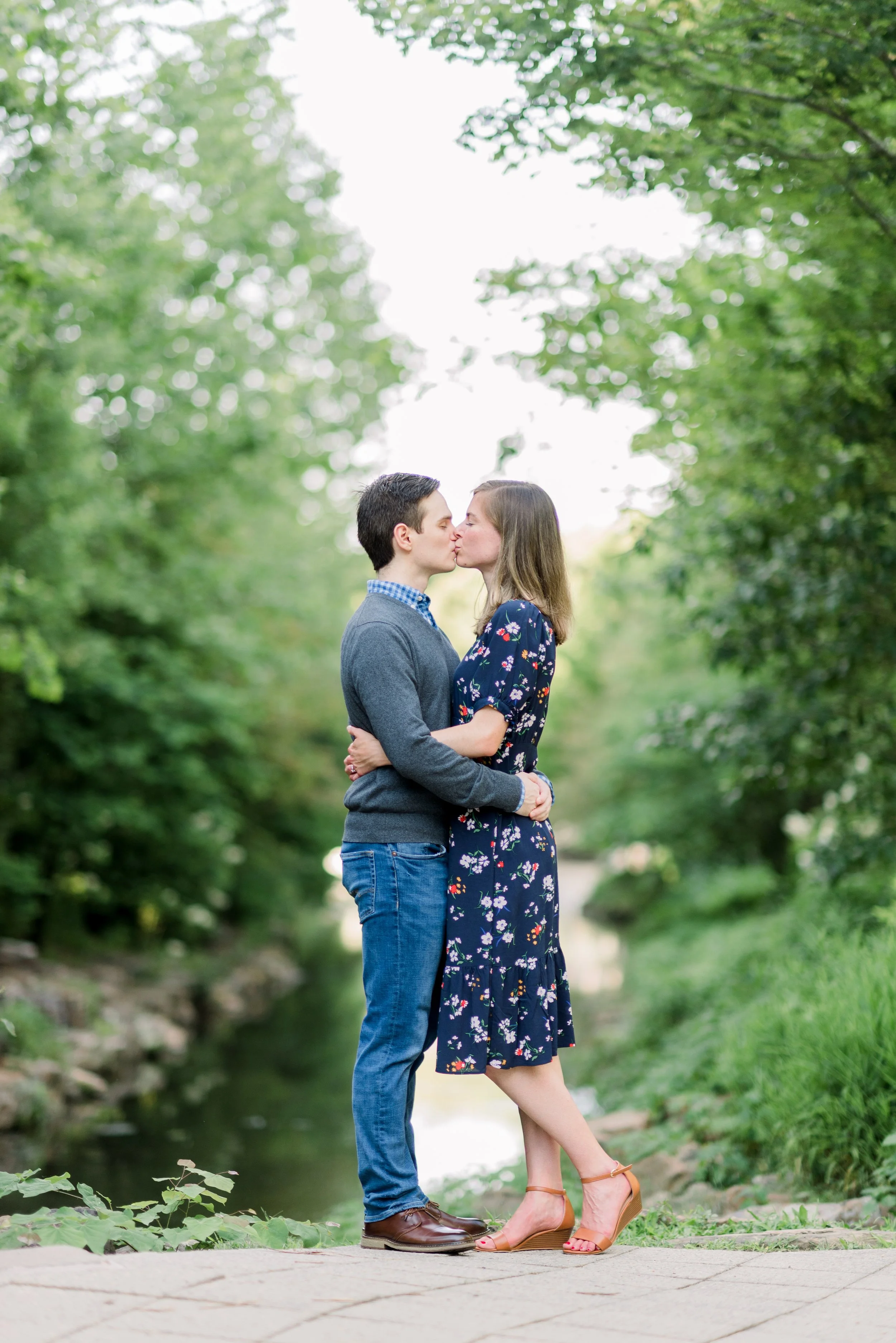 A couple kissing outdoors near a stream, surrounded by lush green trees in a park or nature setting.