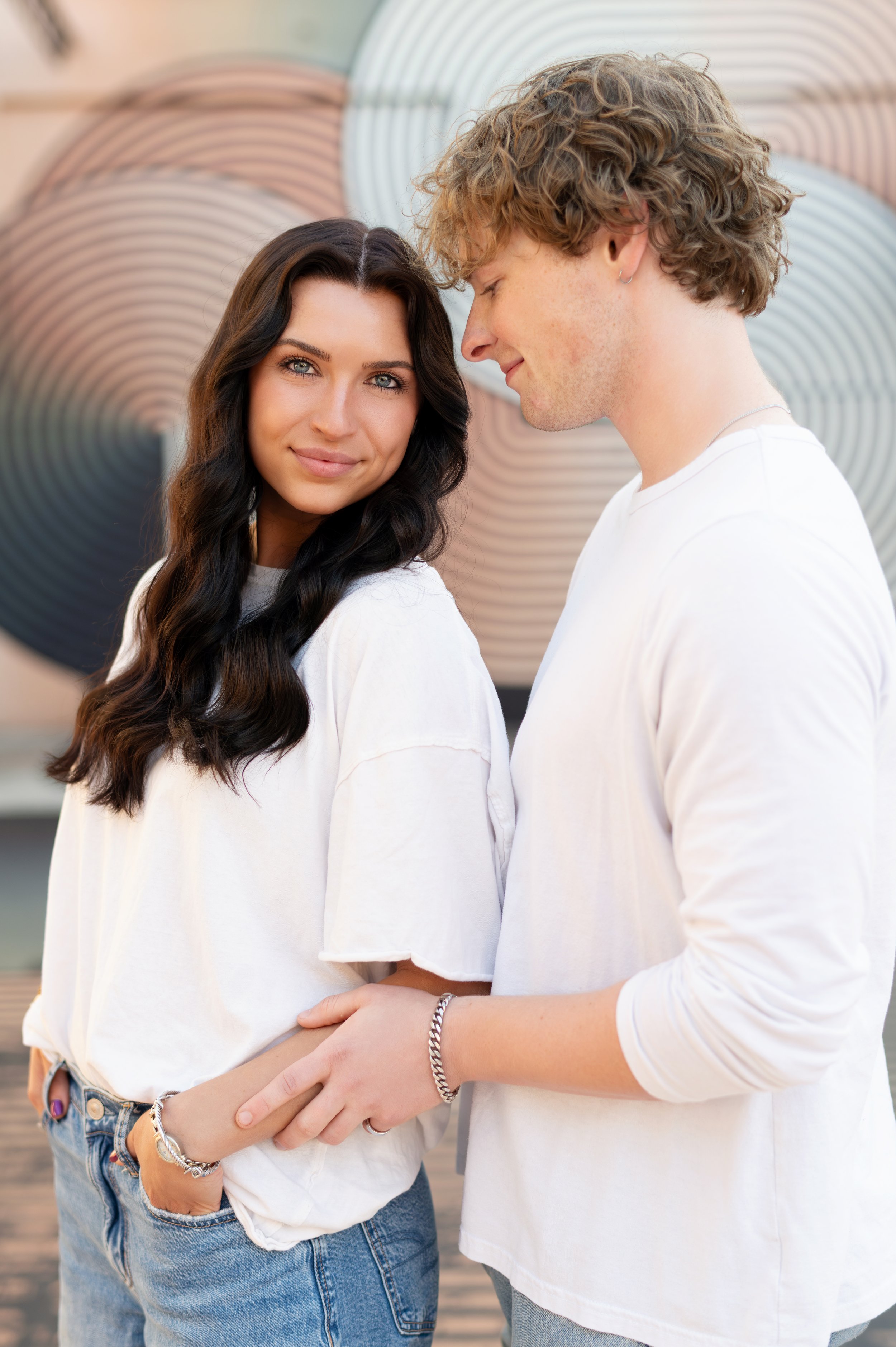 A young couple holding hands and standing close together, with the woman looking at the camera and smiling, and the man with his eyes closed, leaning towards her, against an abstract background with circular patterns.