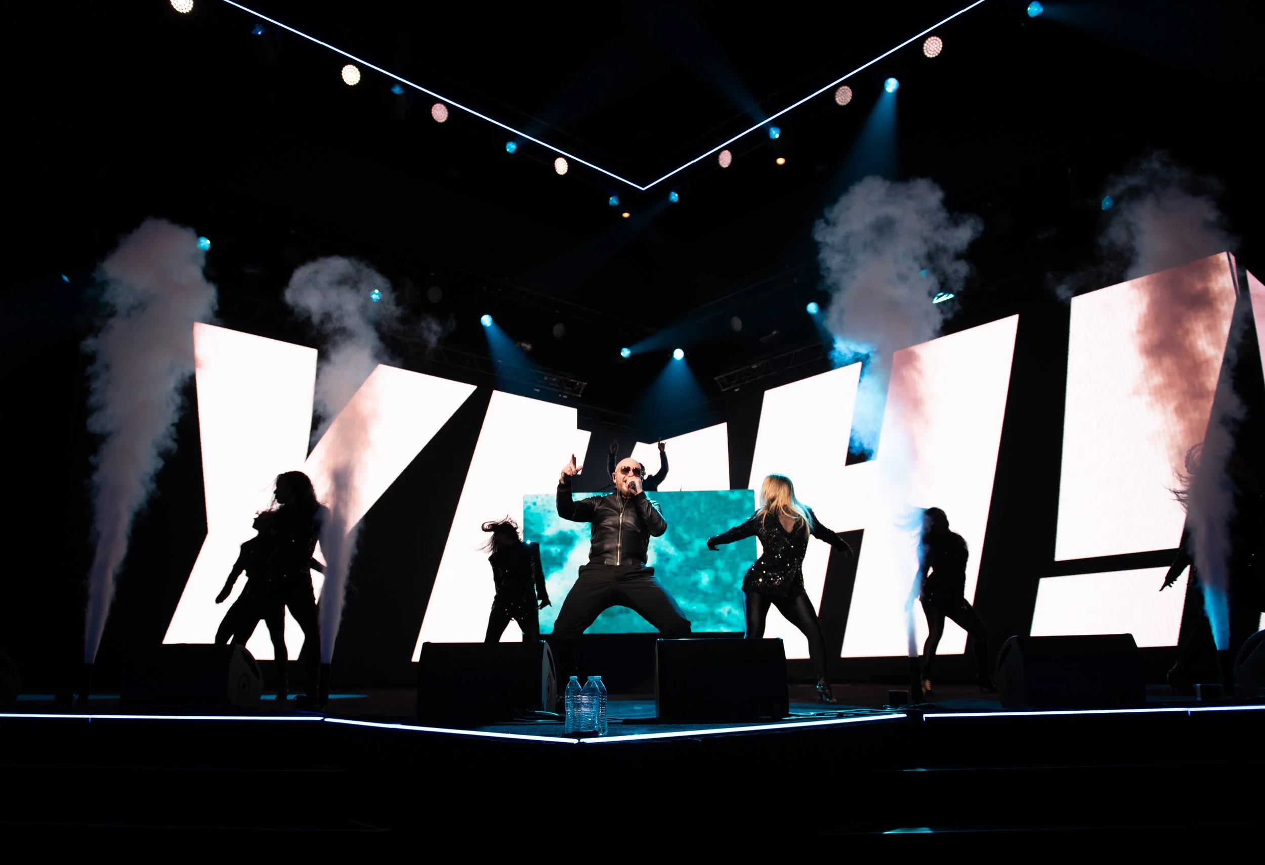 A performer wearing sunglasses and black clothing singing on stage with four dancers in black outfits, with large LED screens and smoke effects in the background.