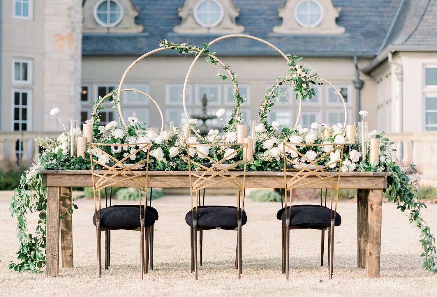 An outdoor wedding table decorated with white flowers, greenery, and gold circular arches, with four gold chairs with black cushions in front, set against a large house with a stone and cream exterior.
