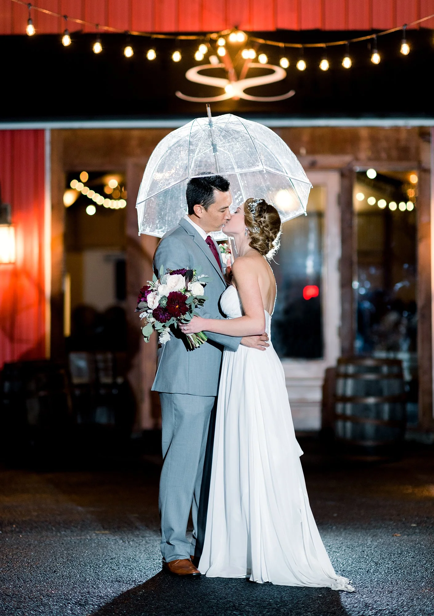 A bride and groom kiss under a transparent umbrella at night. The bride holds a bouquet of dark red, white, and green flowers, and they are standing in front of a warmly lit venue with string lights.