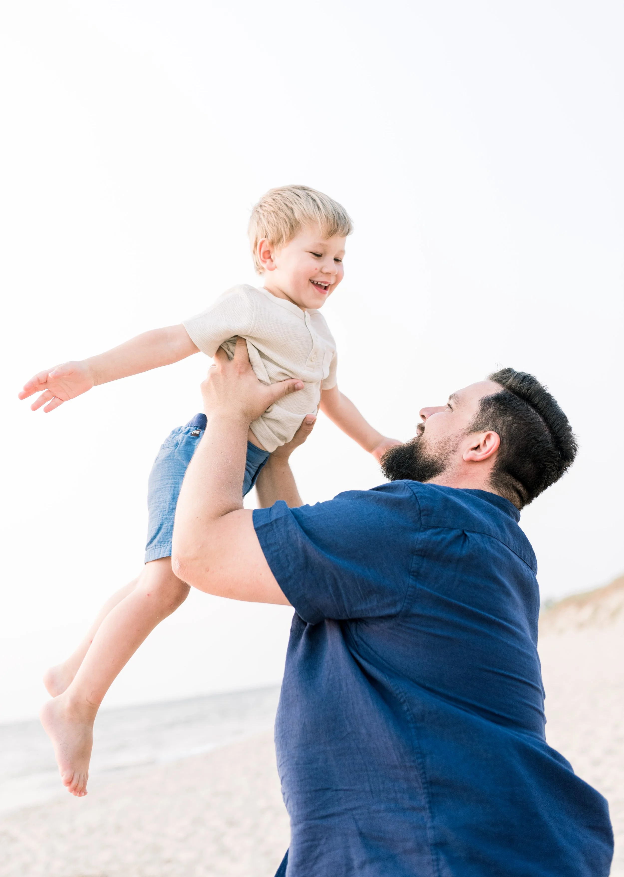 A man with a beard holding a smiling young boy in the air at the beach.