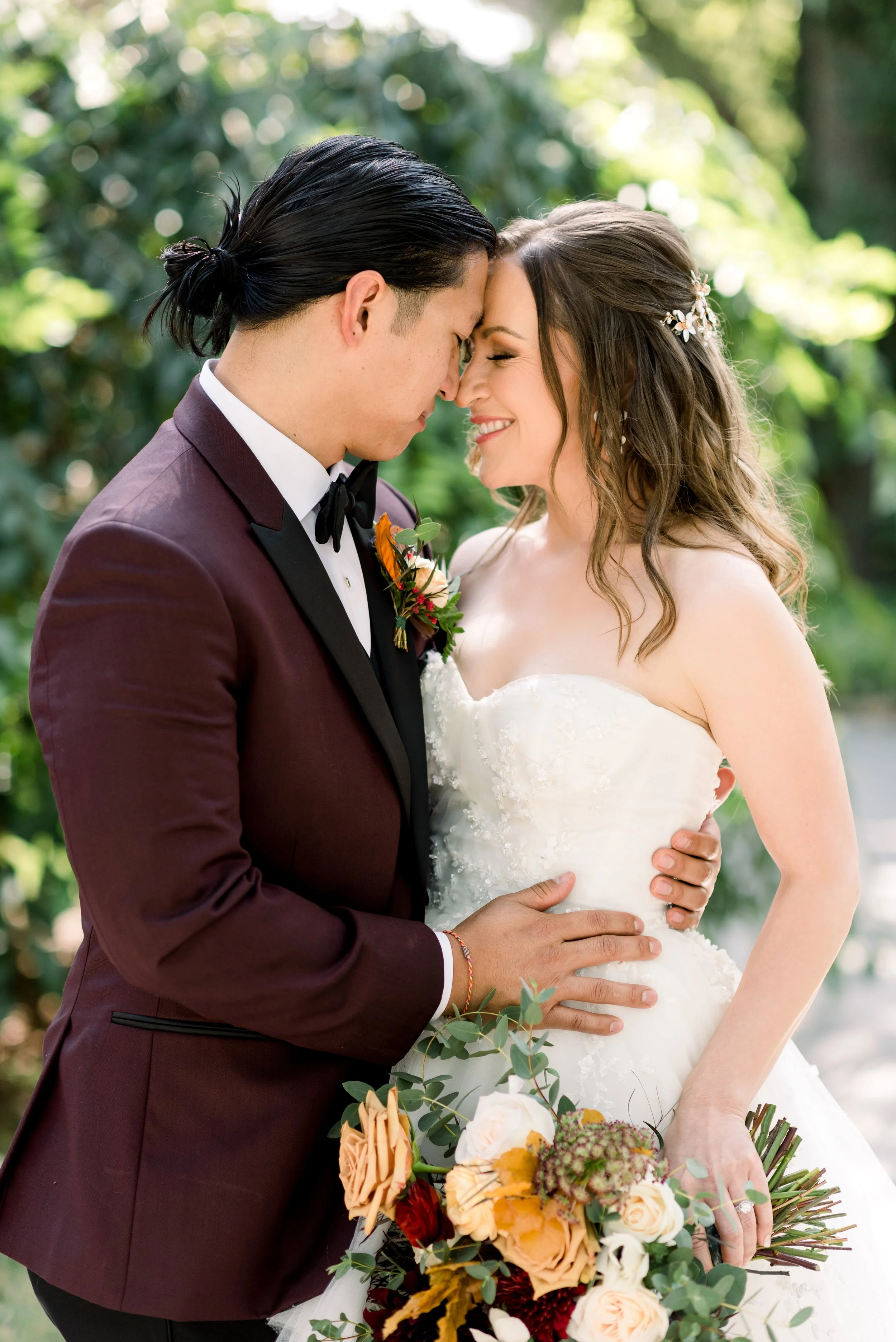 A bride and groom holding each other closely with foreheads touching outdoors, surrounded by greenery, with the bride holding a colorful bouquet of flowers.