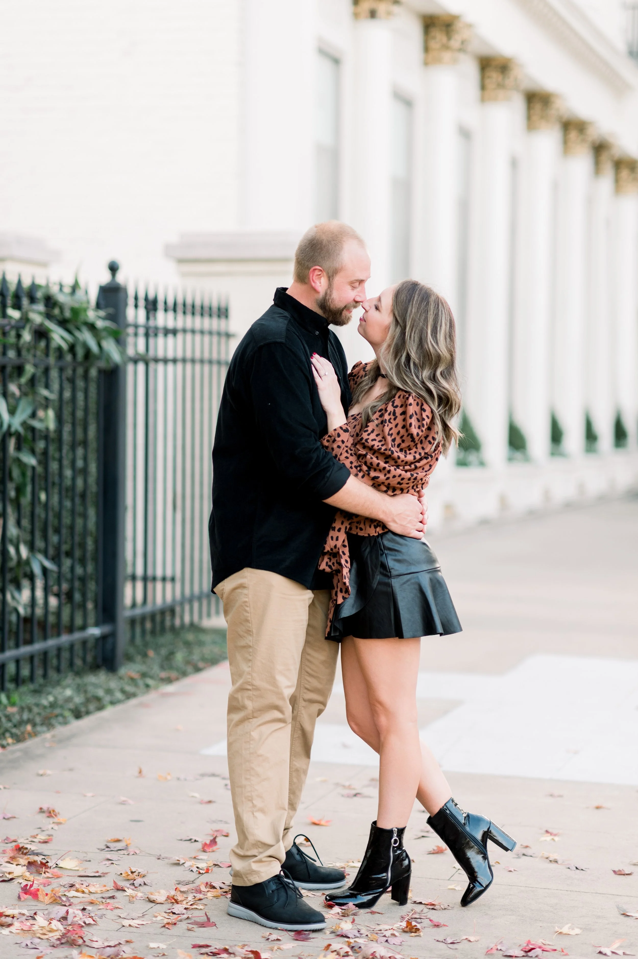 A couple standing close together outdoors on a sidewalk, excitedly leaning in for a nose-to-nose moment with their foreheads touching, surrounded by fallen autumn leaves.