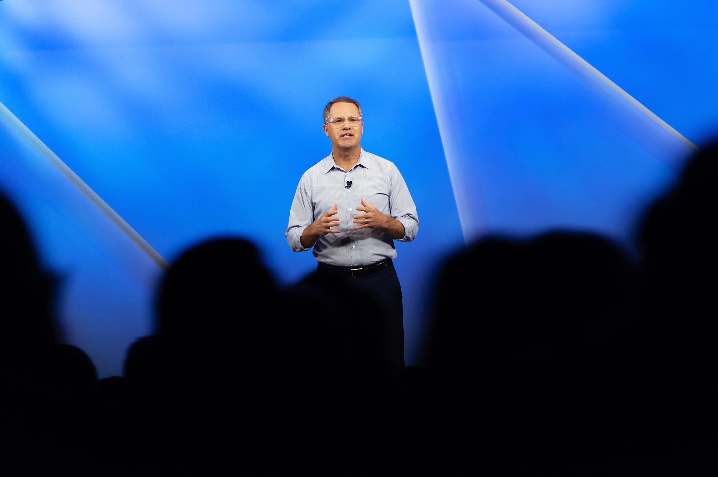 A man in a light blue shirt giving a presentation on stage with a blue background, seen from the audience perspective with silhouettes of people in the foreground.