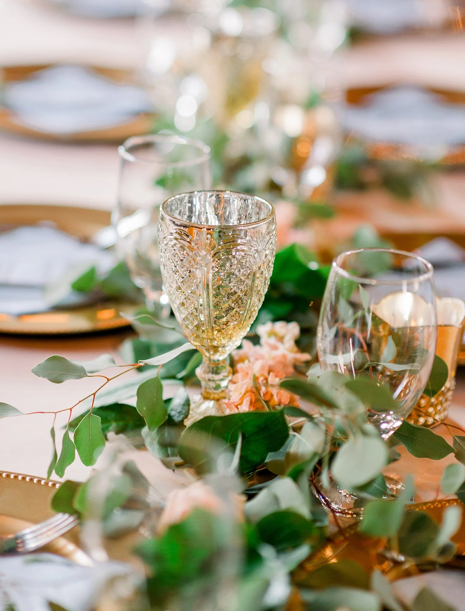 Close-up of a table setting with ornate glasses, greenery, and pink flowers for a formal event or celebration.