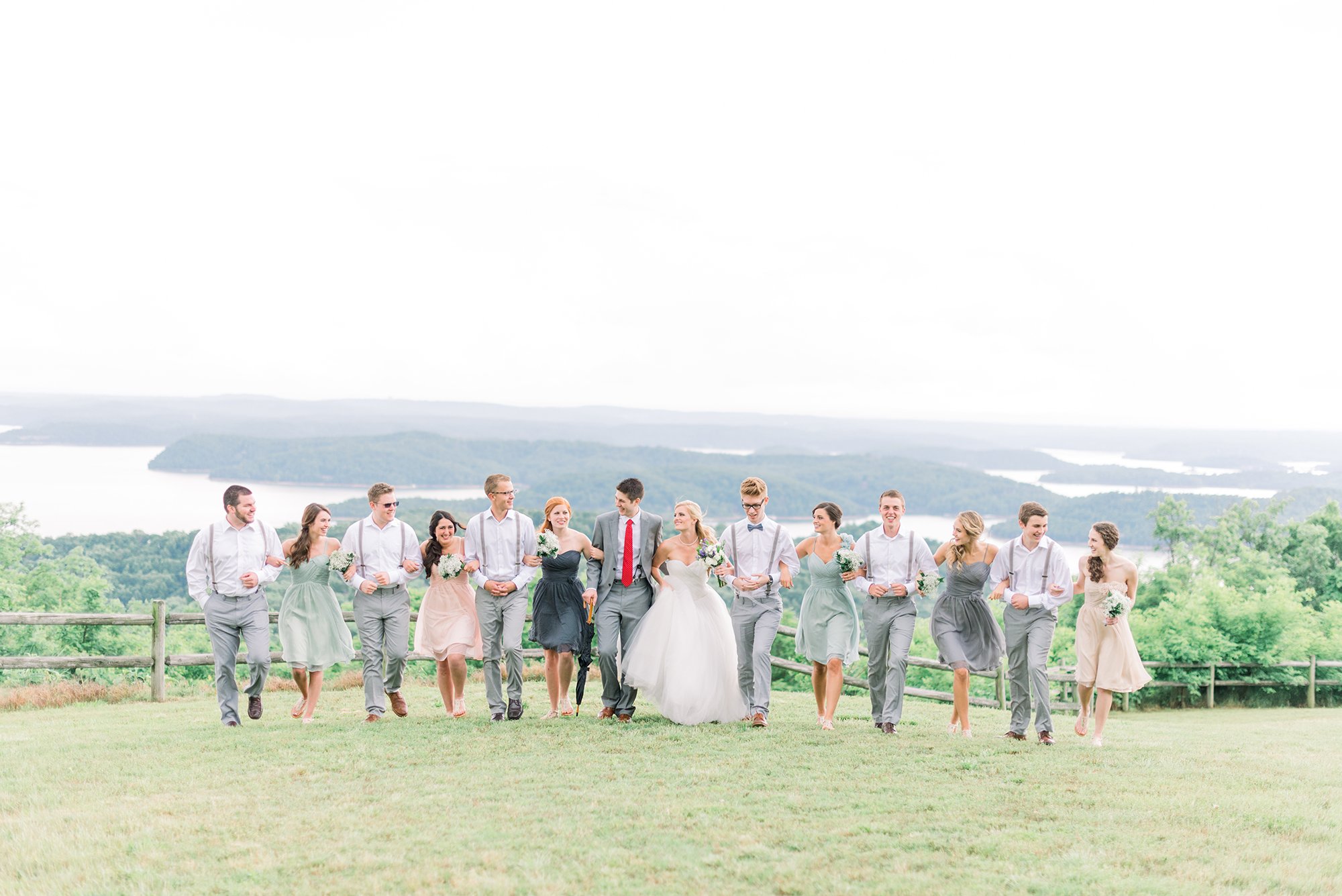 Wedding party walking outdoors on a grassy field with scenic lake and mountains in the background, including the bride, groom, and bridesmaids and groomsmen.