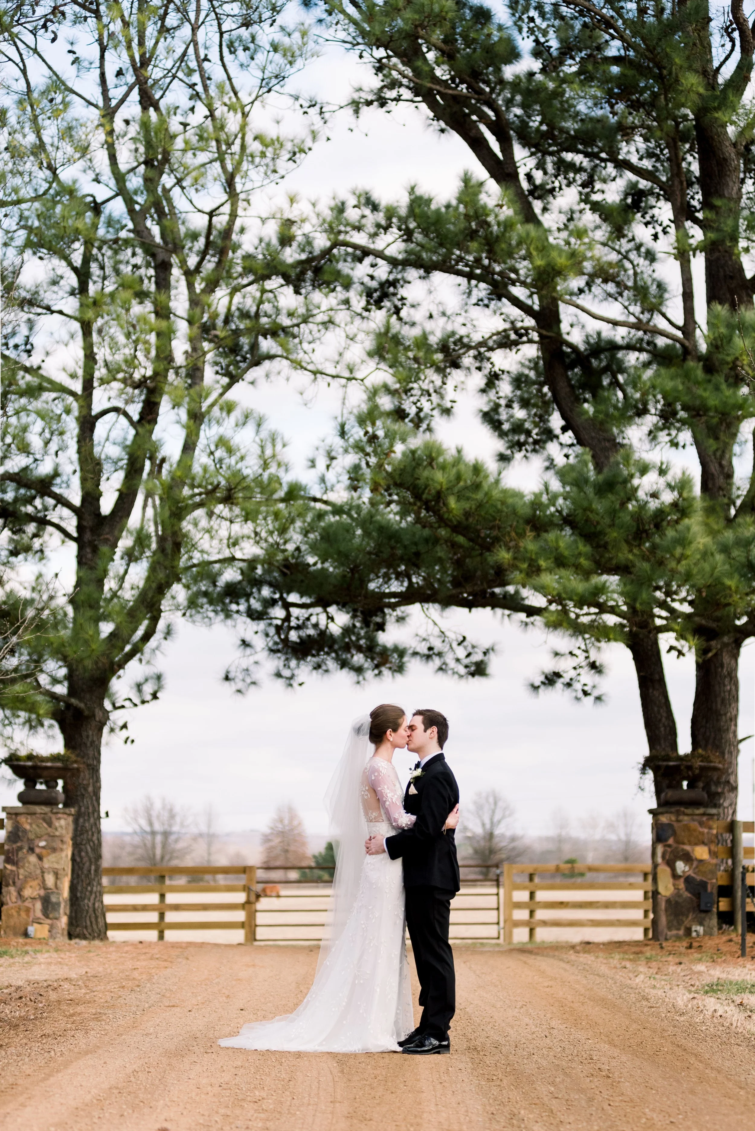 A bride and groom kiss under large trees on a dirt path with a wooden fence and open landscape in the background.