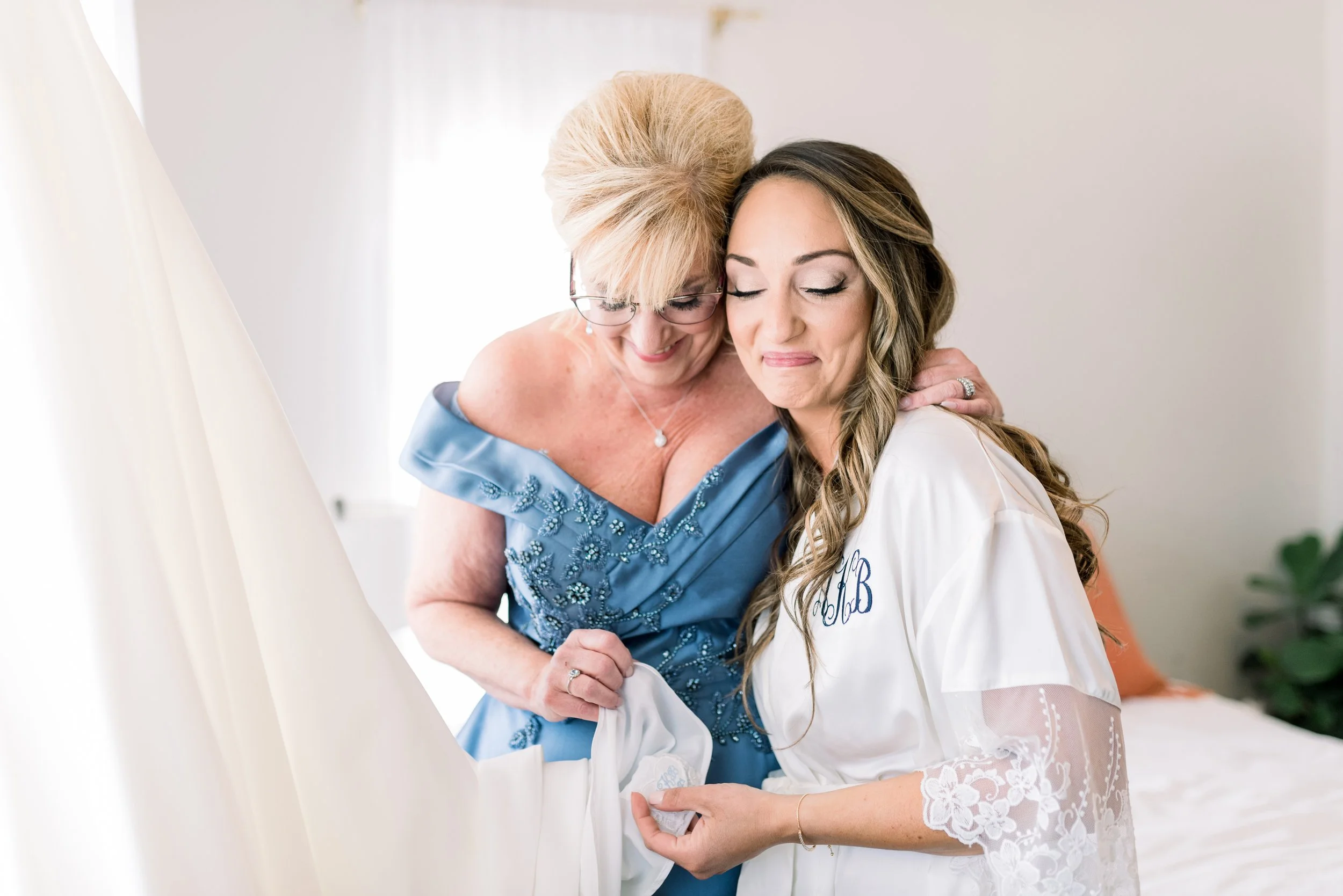 A mother and daughter sharing an emotional moment before a wedding, with the daughter dressed in a white robe and the mother in a blue dress, both smiling with eyes closed.