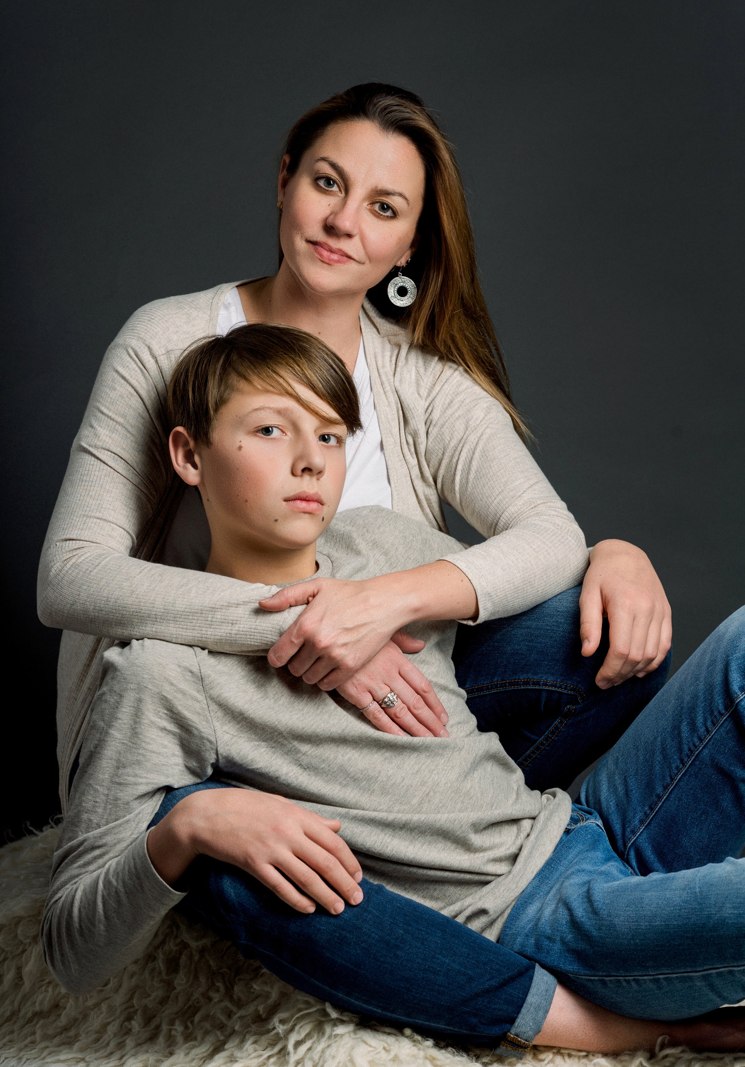 A woman with long brown hair wearing a beige cardigan and earrings, sitting behind a teenage boy with short light brown hair, wearing a gray long-sleeve shirt and jeans, both posing closely together against a dark background.