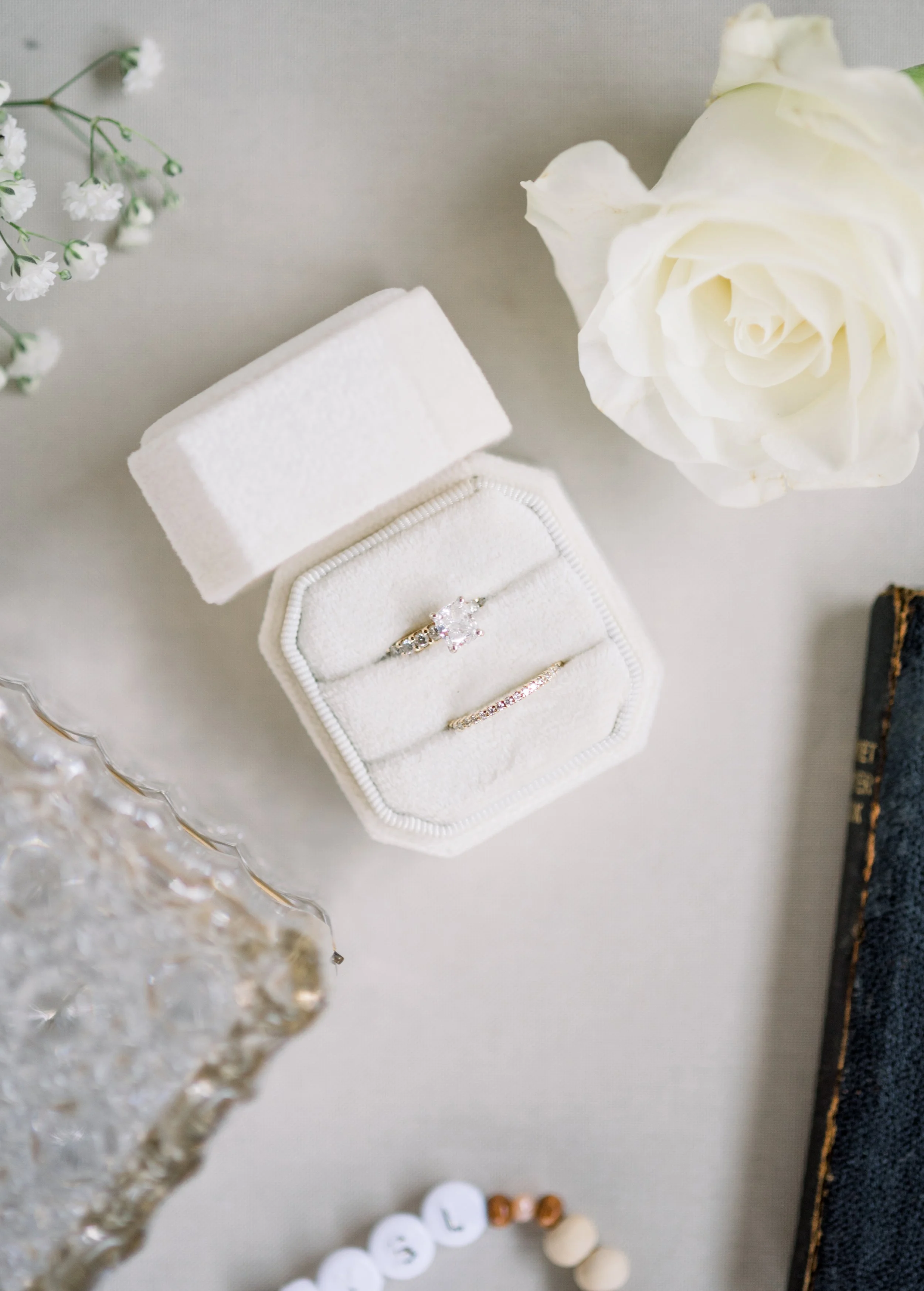 A white ring box with two rings inside, one with a large gemstone and the other with smaller gemstones, placed on a white surface with a white rose and small white flowers nearby.