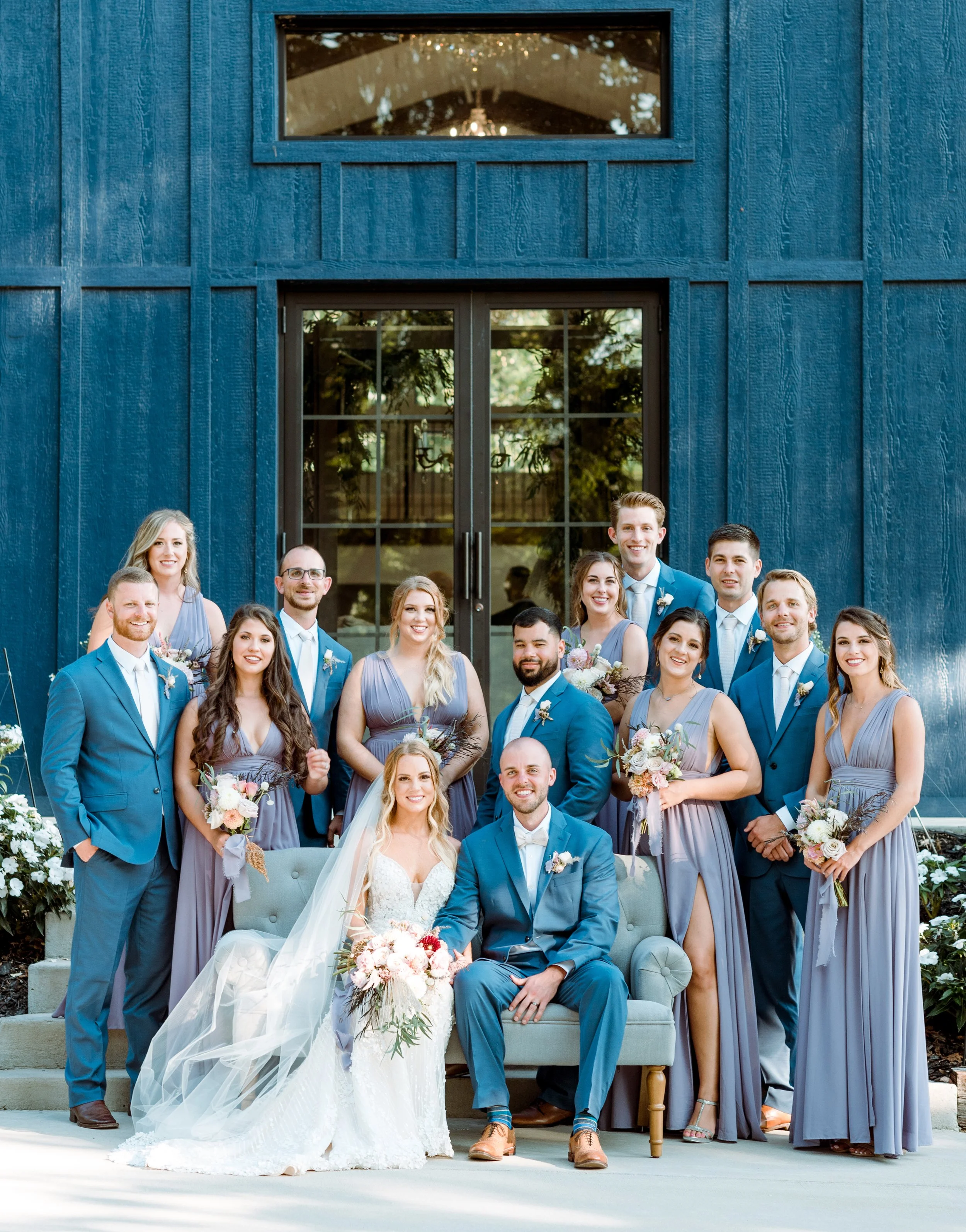 Group of wedding guests, including bride and groom, standing and seated in front of a blue barn on steps, with floral arrangements and dresses in shades of purple.