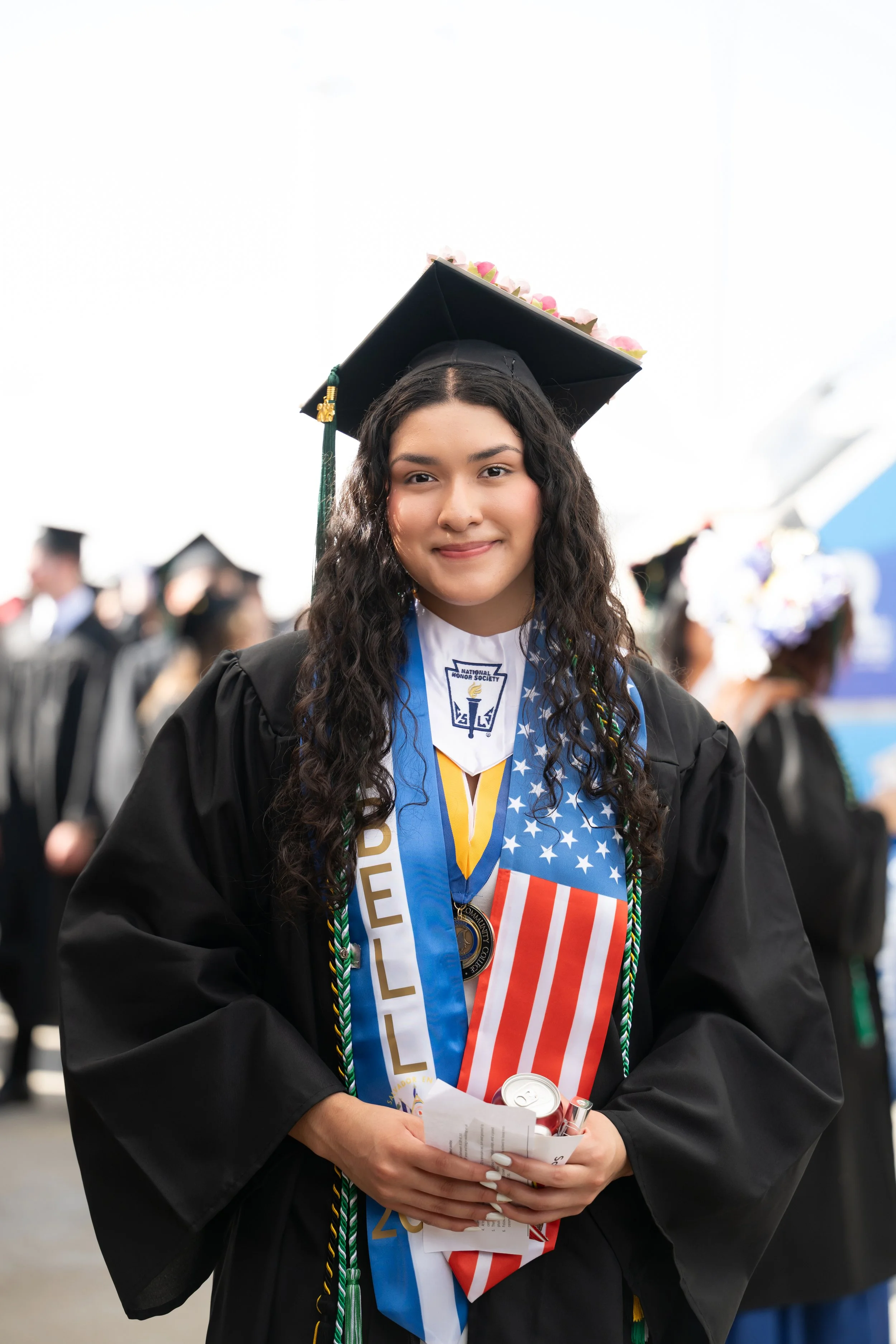 A young woman in a graduation cap and gown at a graduation ceremony, holding a diploma, with a background of other graduates.