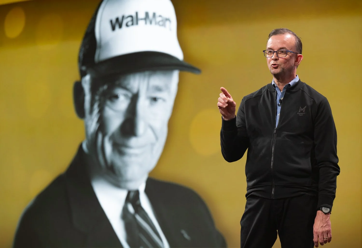 A man wearing glasses and a black jacket is speaking on a stage, pointing with his right hand. Behind him, a large black-and-white image of a person in a suit and a cap with the word 'Walmart' on it is displayed on a yellow background.