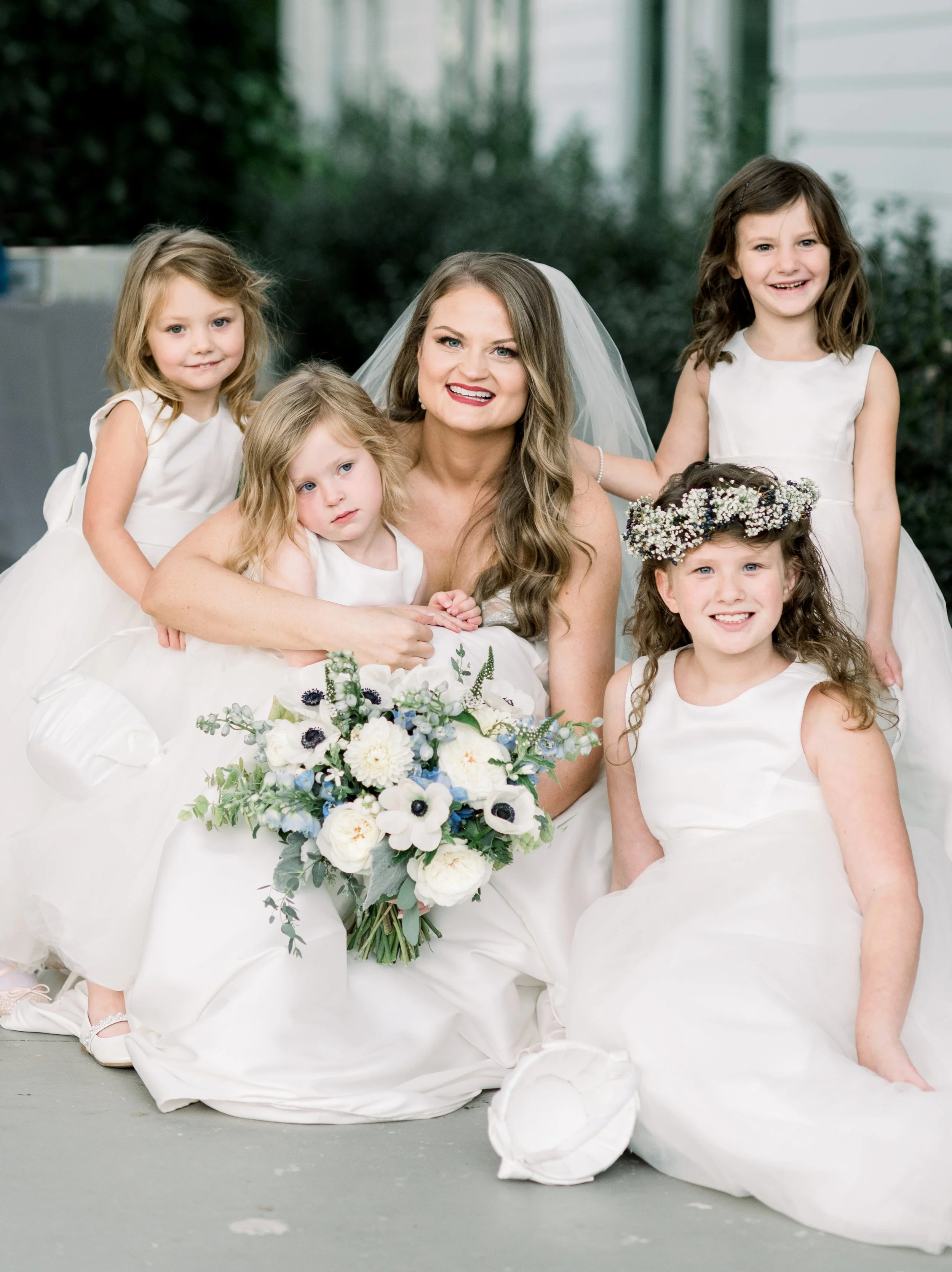 A bride in a wedding dress and veil, holding a bouquet of white flowers and greenery, surrounded by five young girls in white dresses, some wearing floral headbands, outdoors with greenery and a house in the background.