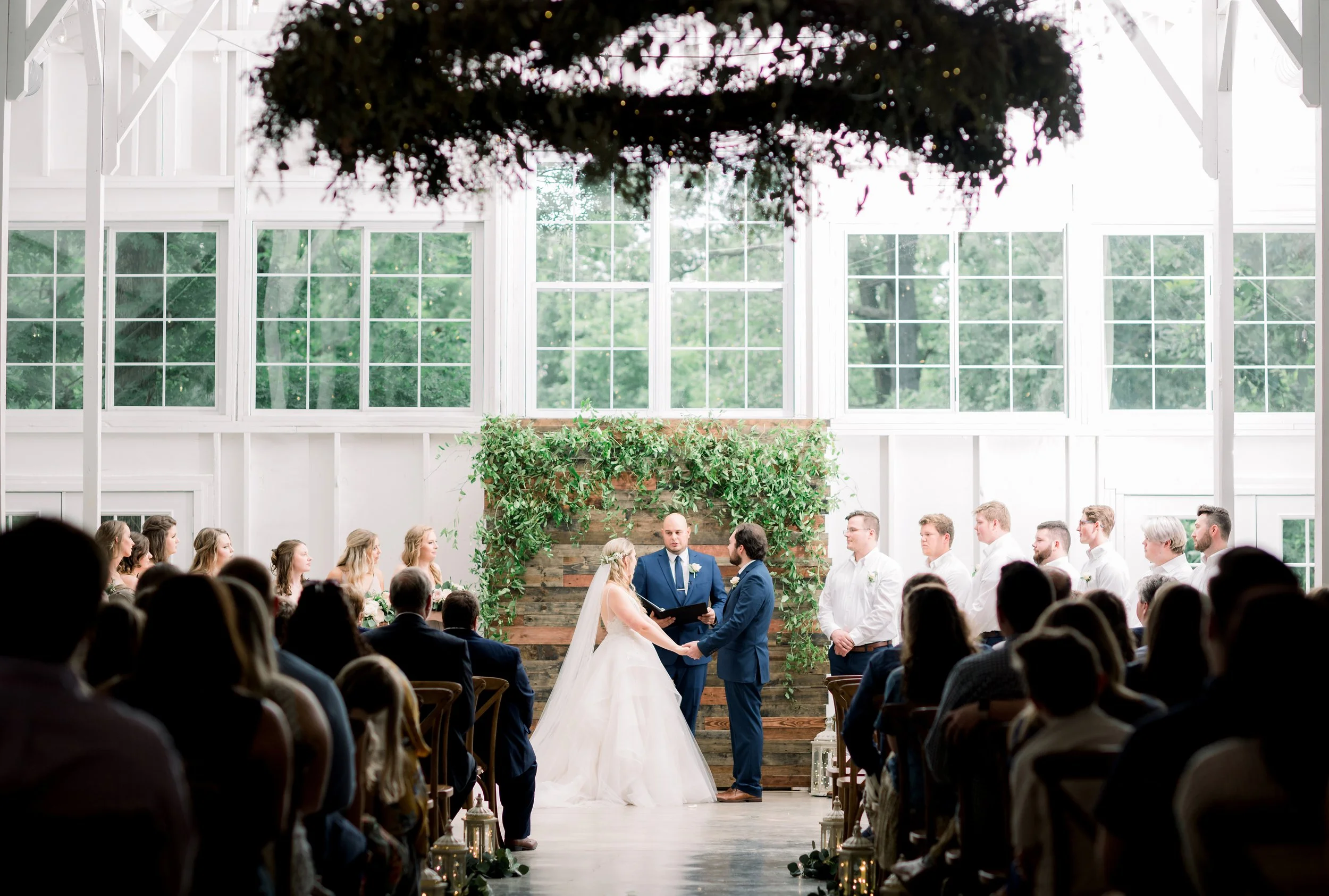 Indoor wedding ceremony with a bride and groom holding hands in front of an officiant, surrounded by guests seated in rows. The background features large windows and a decorated wooden backdrop with greenery.